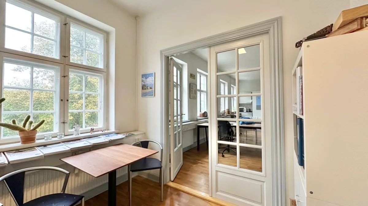 Inde i et studierum med trægulv og højt loft står et bord og to stole med udsigt til haven