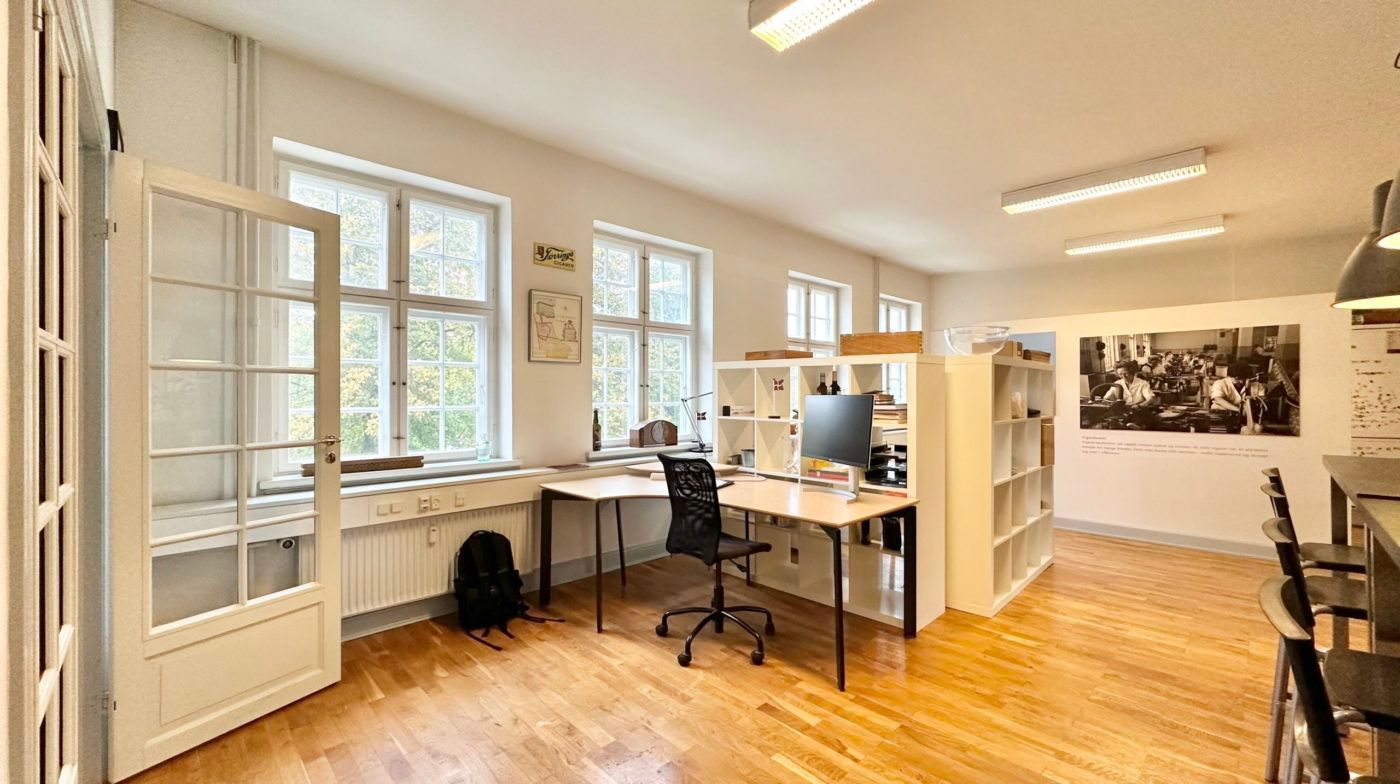 Et lyst kontor med skrivebord og stol i en af siderne, et højt hyldesystem og flere lysekroner på loftet