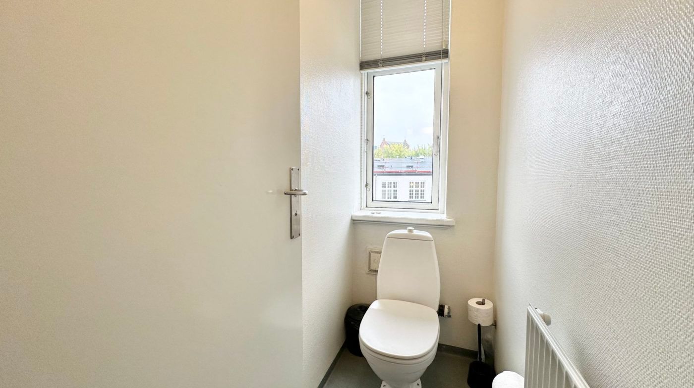 En lille toiletrum med hvid toilet, toiletpapir, hængetoilet og renseri. Der er et vindue med gardiner og udgangsdør med