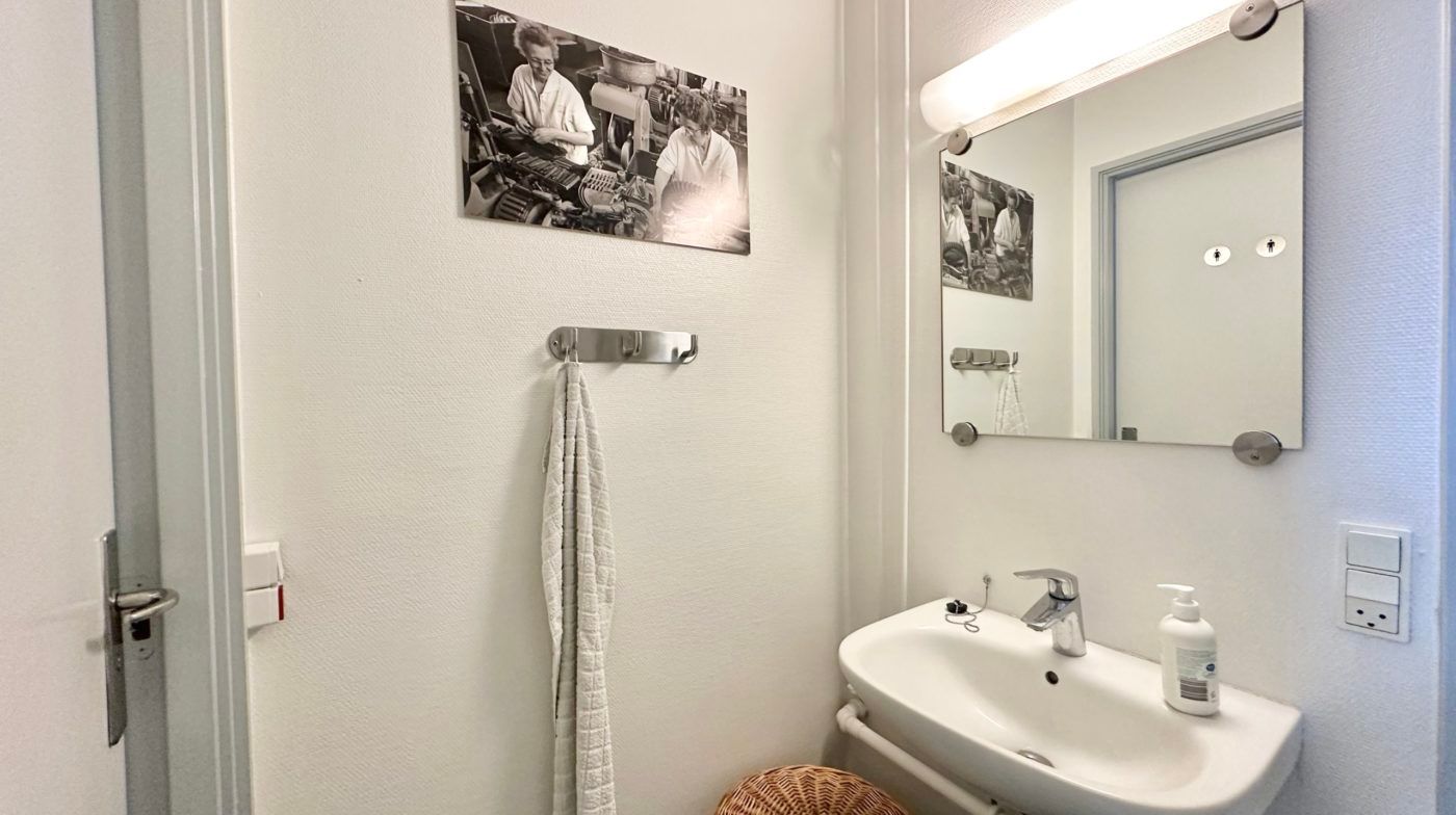 Et rent badeværelse med hvidt porcelænsbassin og håndklædehænger, en spejl med lysearmatur, og en sæbeflaske samt