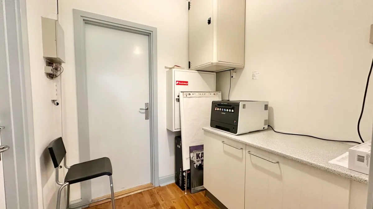 En lille stue med hvid væg og trægulv. På loftet er der en printer. En kæmpe hvid kabinetsstruktur er monteret på væggen. En