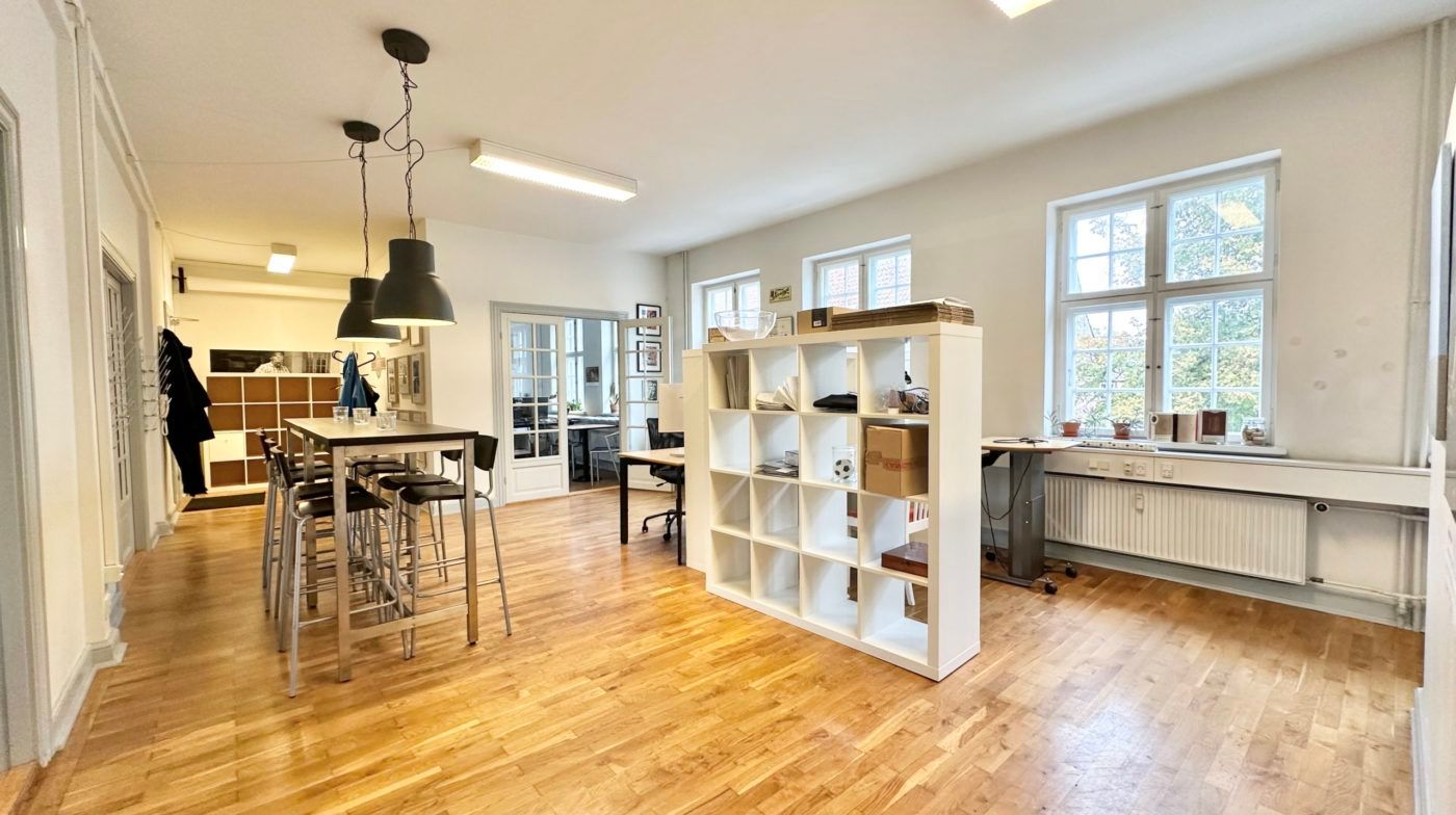 Lyst og ryddeligt kontor med højloft, lyseruder og trægulv, højt opstillet hvid hylde og bord med stolpe og fodstøtter