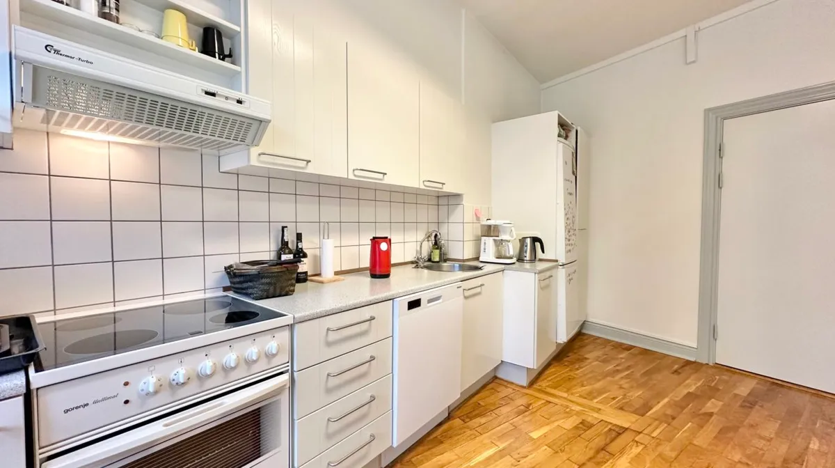 Et tørrehylde med lysegul tønde og sort kaffemaskine. Et rent køkken med gaskomfur og ovn. Et hvidt køleskab og et bord med