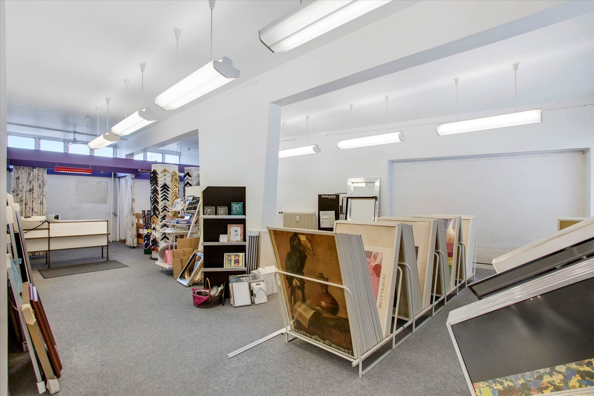 Godt oplyst lokale med lyse gulve og vægge, hængende lys og hylder med kunstværker og indrammede billeder samt