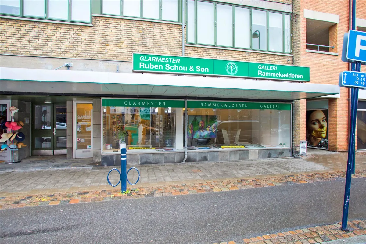 Synlig butik med navnet Glarmeister Ruben Schou & Søn, store facadevinduer, og udsmykning på siderne af butikken med