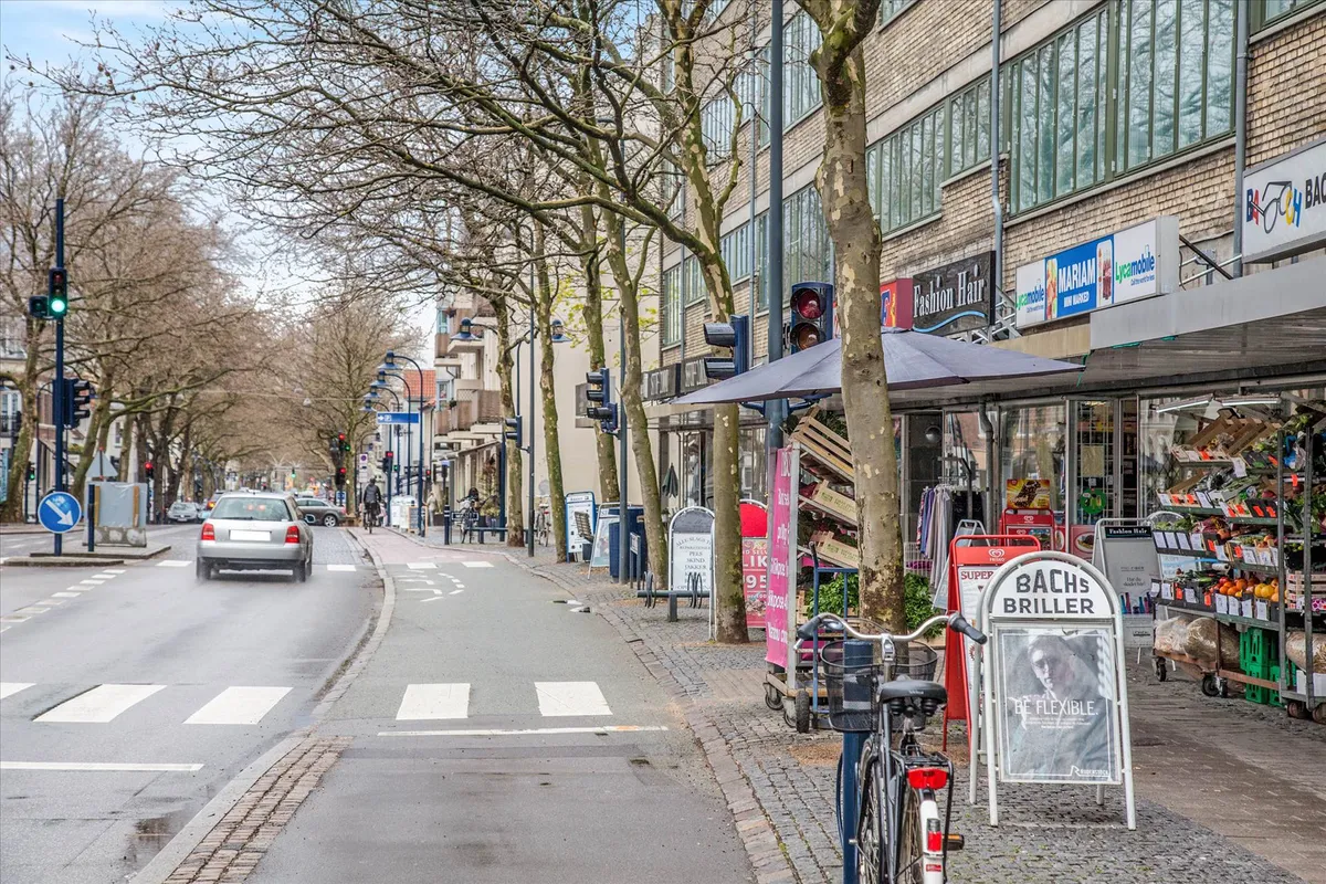 Cykelparkering udenfor Bachs Briller med friske frugter og grøntsager og et bil på vejen med forskelligt trafiksignal