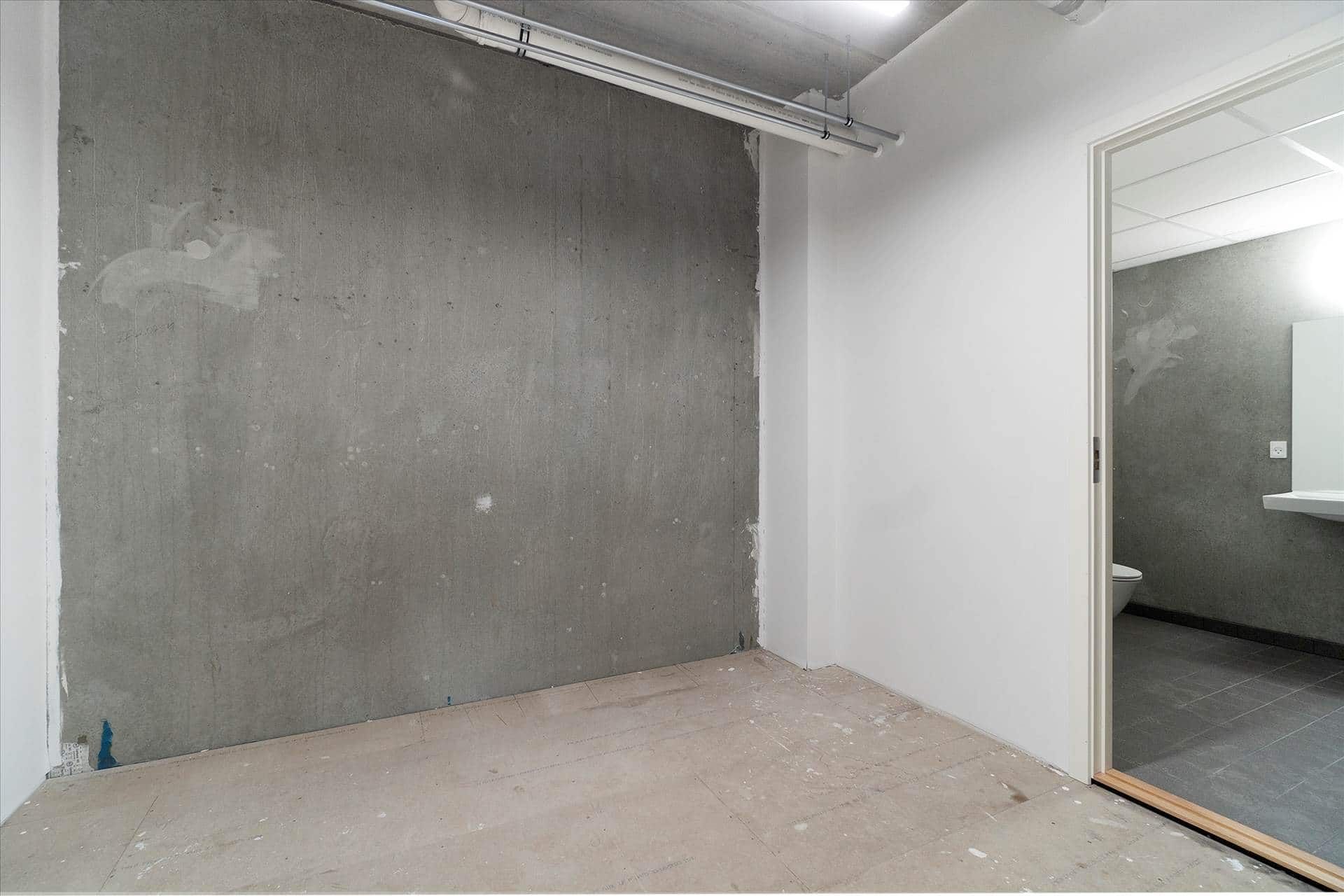 En tom rum med ubehandlet betonvæg og lysegrå gulv, hvor døren til badeværelset er åben og viser toilettet