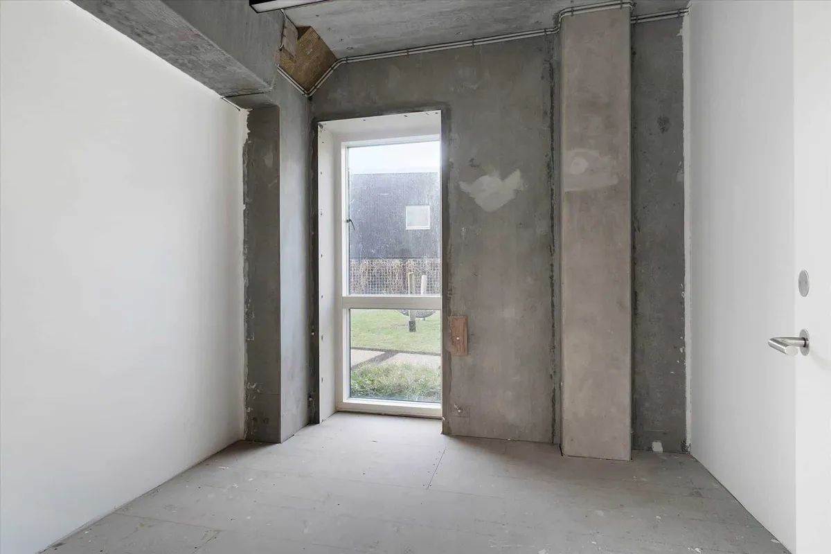 Et ubeboet rum med krydsfinerbelagt gulv og hvide vægge og døre samt et stort vinduesparti med udsigt til et græsfelt