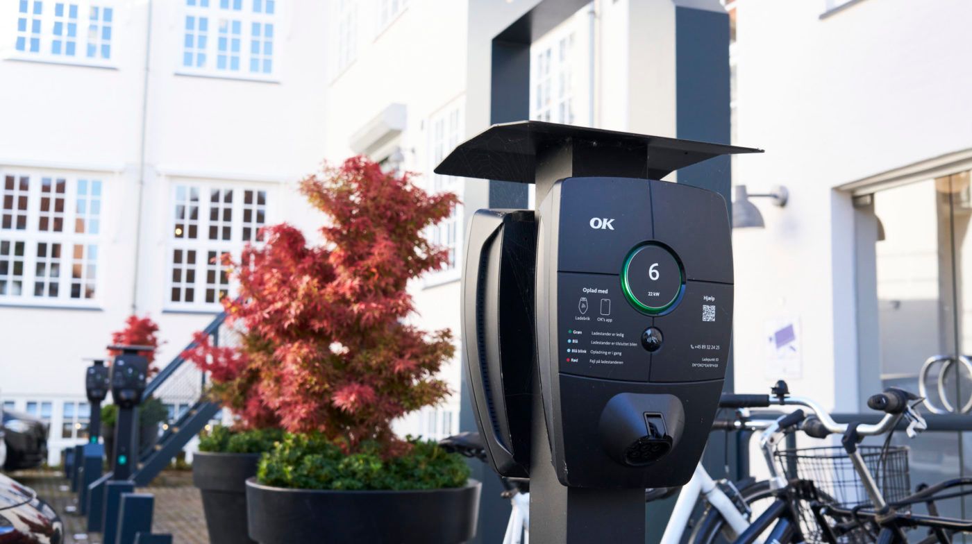Ladestation til elbiler med OK-logo og en grøn cirkel med tal 6, to cykler parkeret og to buske med rød og grøn blade