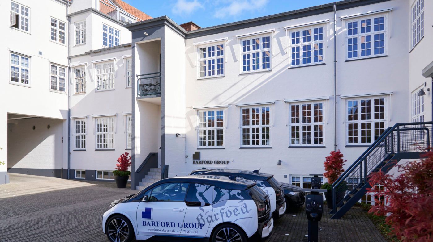 En hvid bygning med Barfoed Group skrift på facaden og to biler parkeret foran og en stigning til hovedindgangen