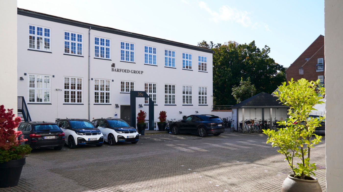 Flere biler er parkeret foran Barfoed Group, der er en hvid bygning med store vinduer og en sort tekst i facaden