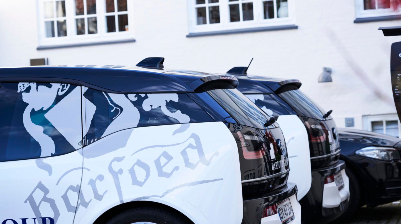 Flere biler parkeret i række foran bygningen. Forreste bil med logo og tekst på siden. Bagved en anden bil med logo på