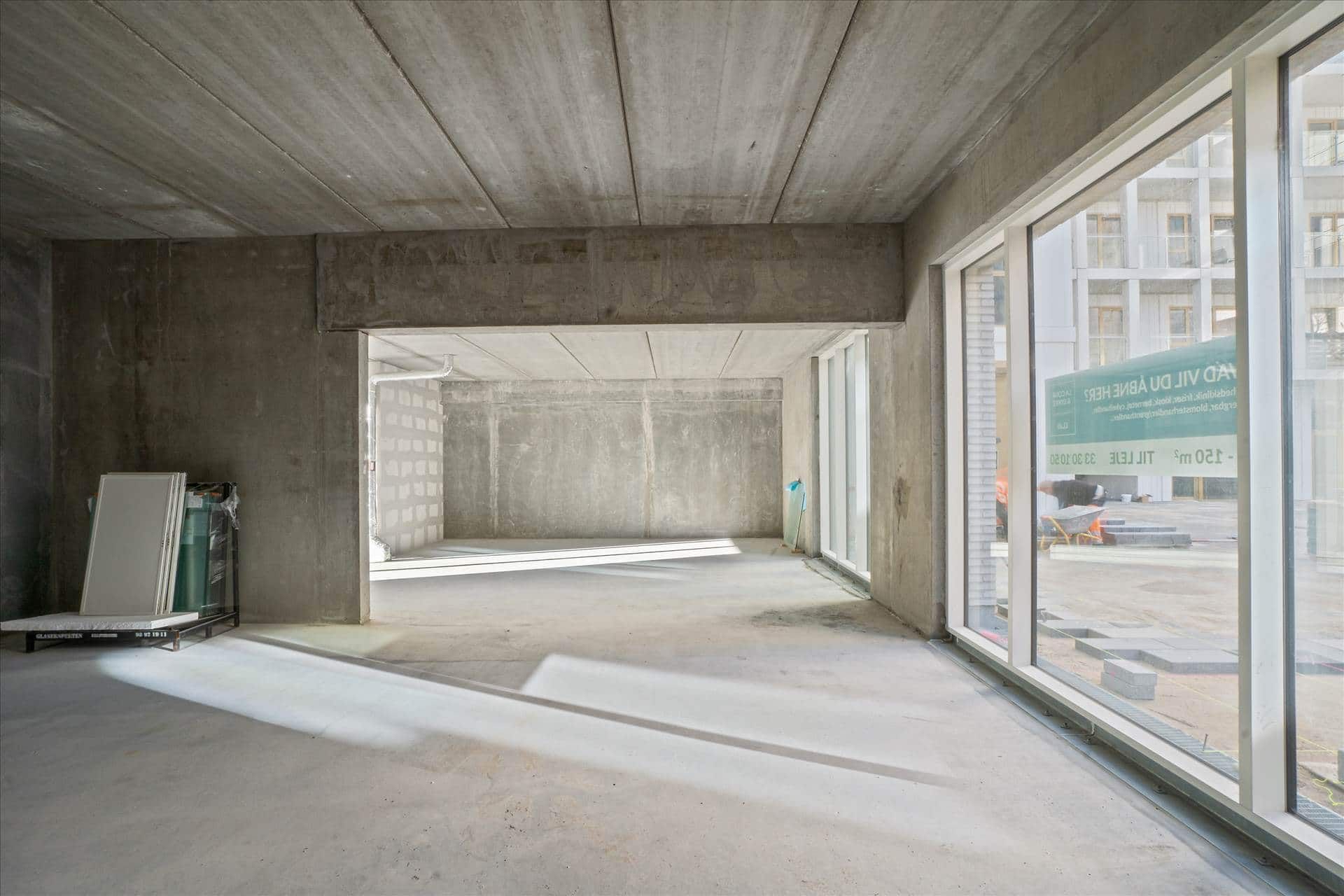 Betongulv og -vægge i et ubeboet rum med to vinduer, der vender ud mod et værkstedsområde med arbejdere og materialer