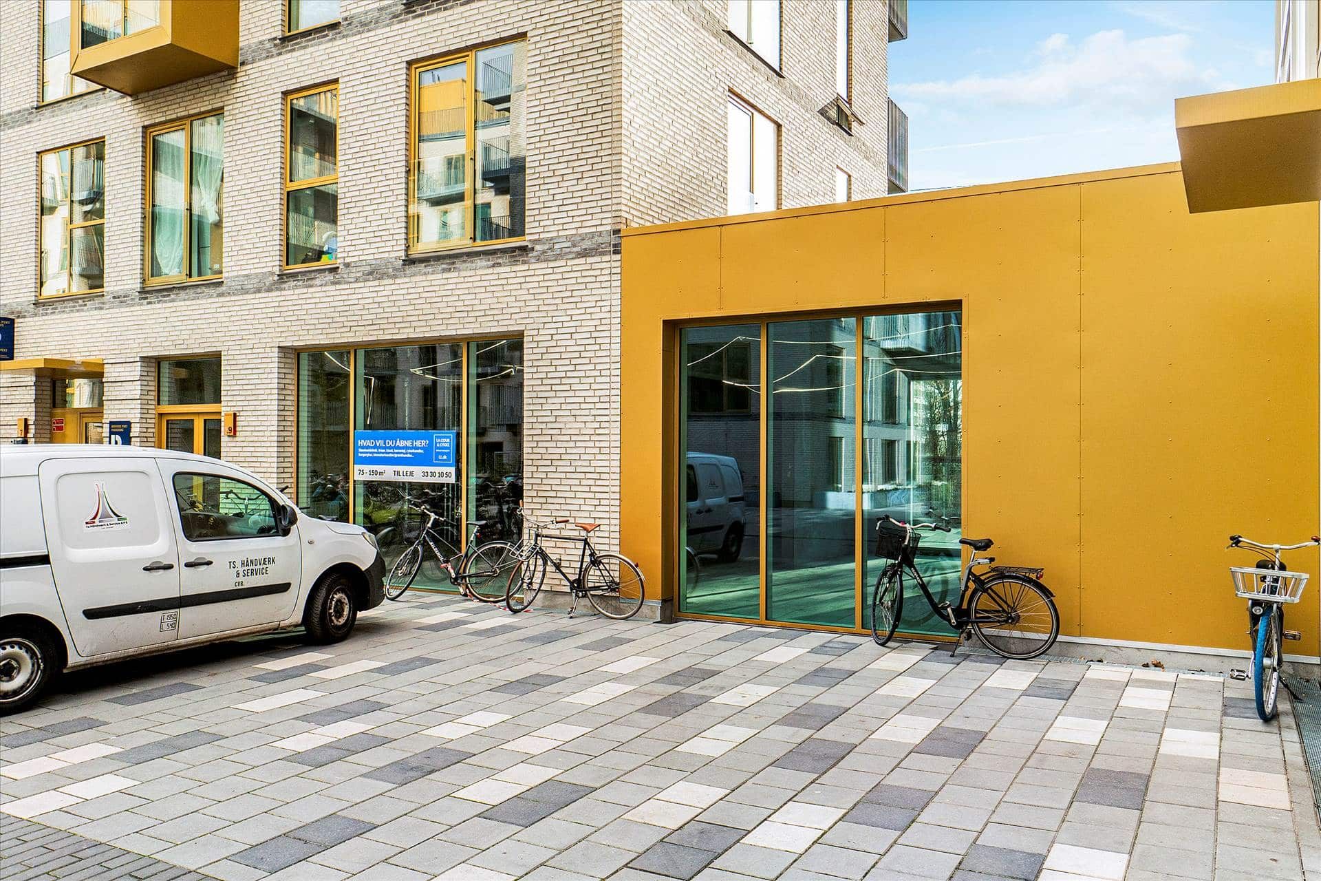 Hvid lastvogn parkeret udenfor butik med gule vægge og glasdøre, flere cykler står parkeret udenfor.