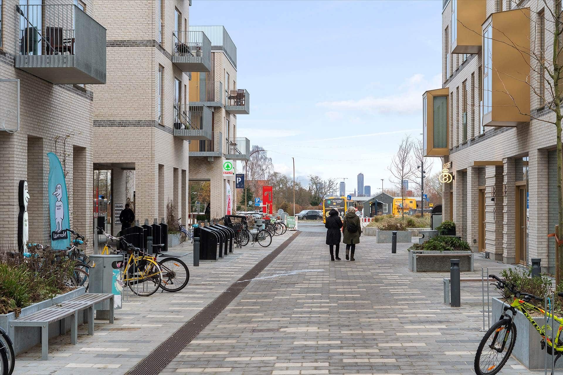 To mennesker går ned ad en gågade med forskellige faciliteter, som cykelstativer og plantager, og en parkeringsplads med