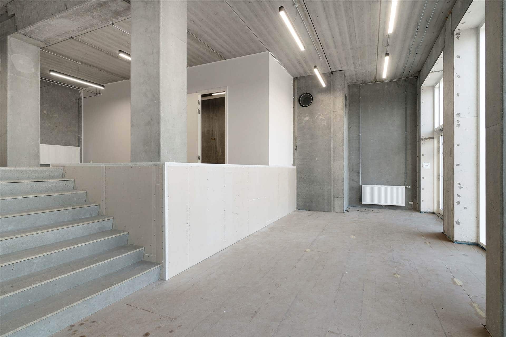Udenfor er der en stor åben erhvervslokal med lyse gulve og lofter. Der er et hjørne med trapper og hvide vægge. Loftet er