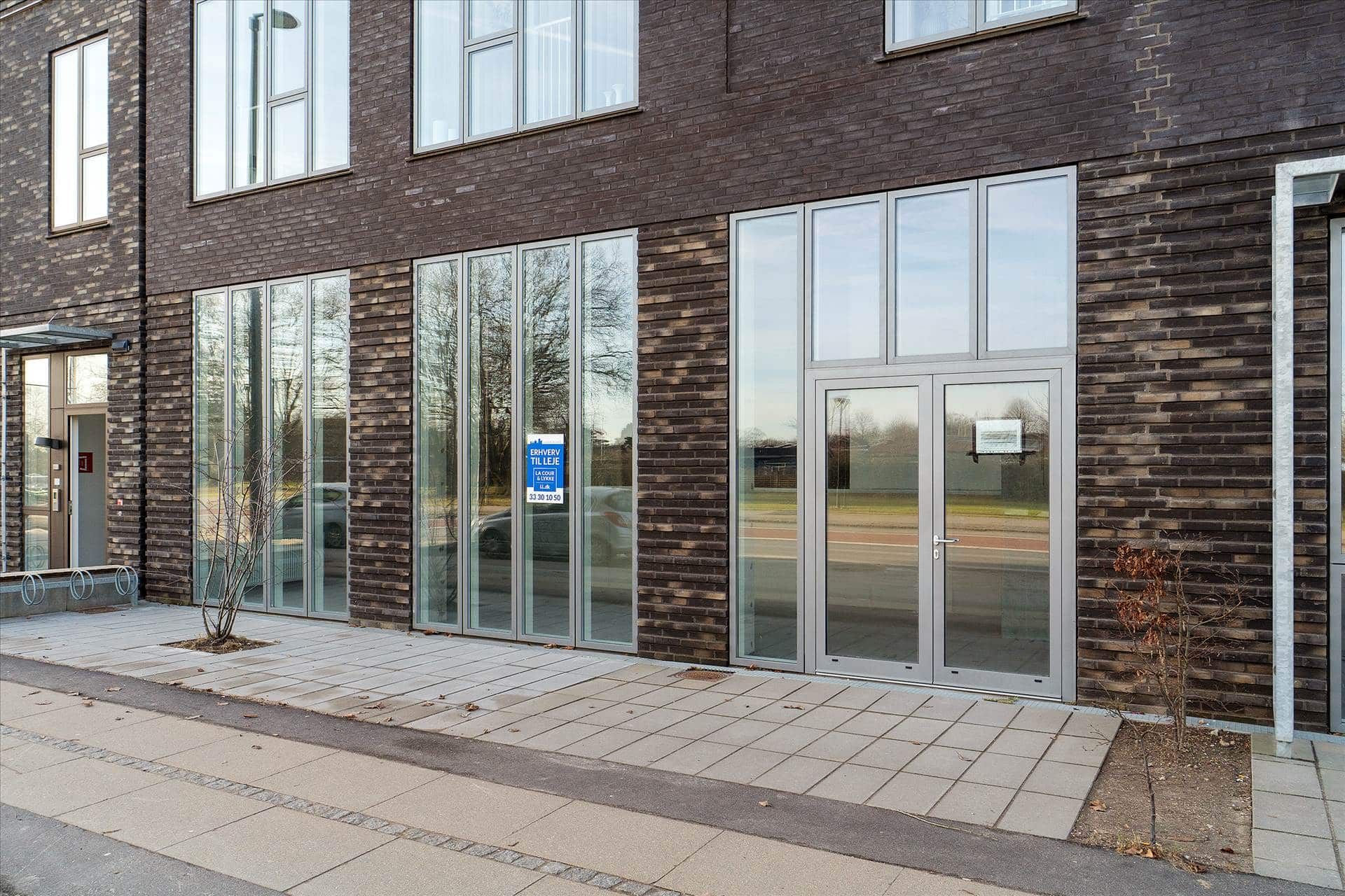 En stor erhvervsbygning med glasvinduer og brun mursten, lyse gulve og en tydelig annonce på vinduet