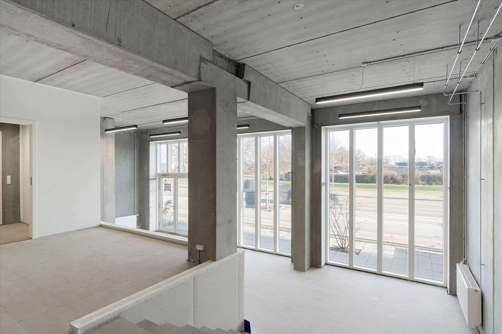 Store, lyse kontorer med hvide vægge, store ruder, lysekroner og trægulve i en bygning med eksponeret beton