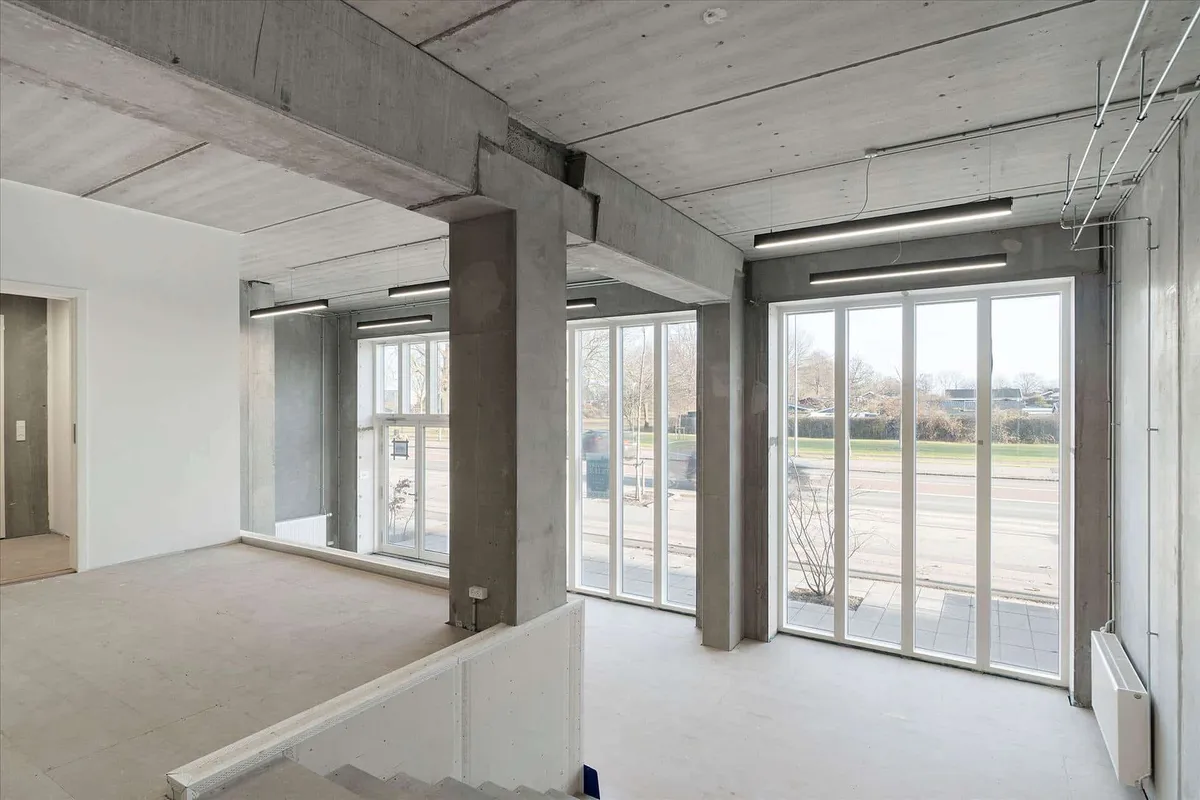 Store, lyse kontorer med hvide vægge, store ruder, lysekroner og trægulve i en bygning med eksponeret beton