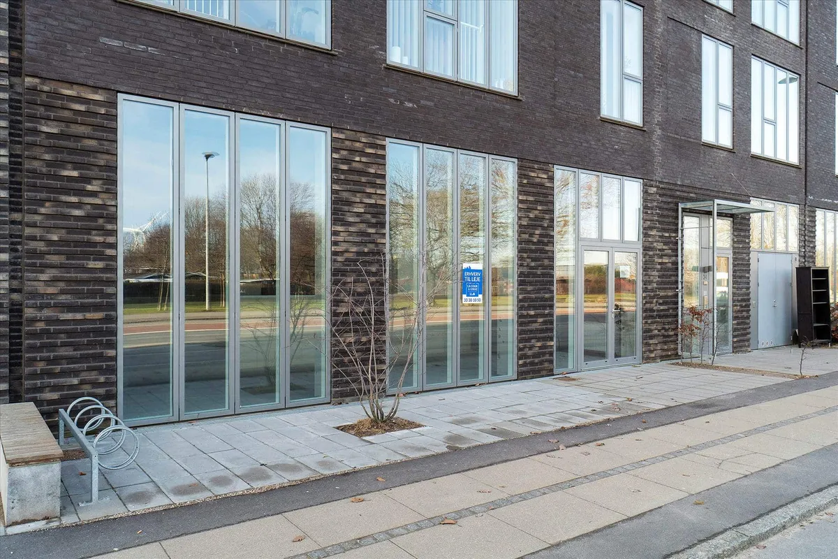 Store, moderne erhvervslokaler i tæt bebygget område med glasvinduer og indgange samt lyse, nye brolægning og plante.