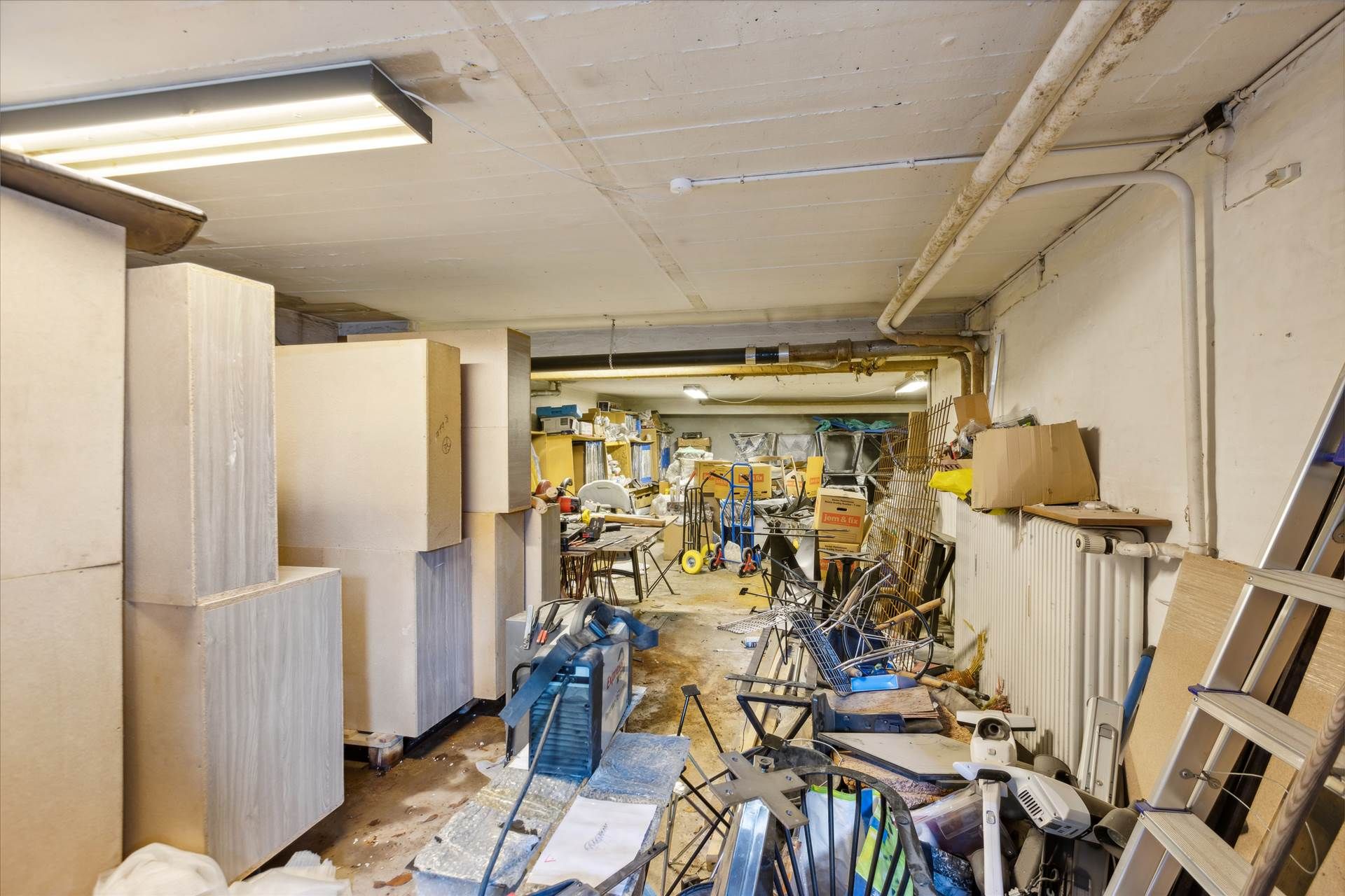 Forladt og rummeligt lager med fysiske skades varer, en bænk, skrald, kasserne og støvsuger på jorden og to lysekroner på