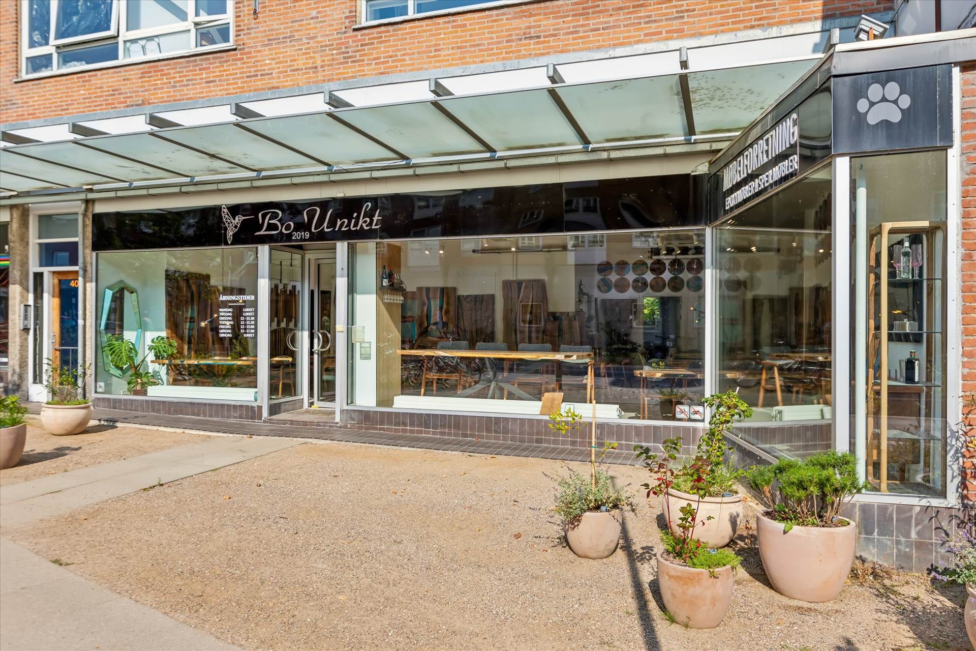 Butiksfront med teksten Bo Uniket og vinduer med plantekrukker og møbler. To store glasruder med spejle og vinduer med