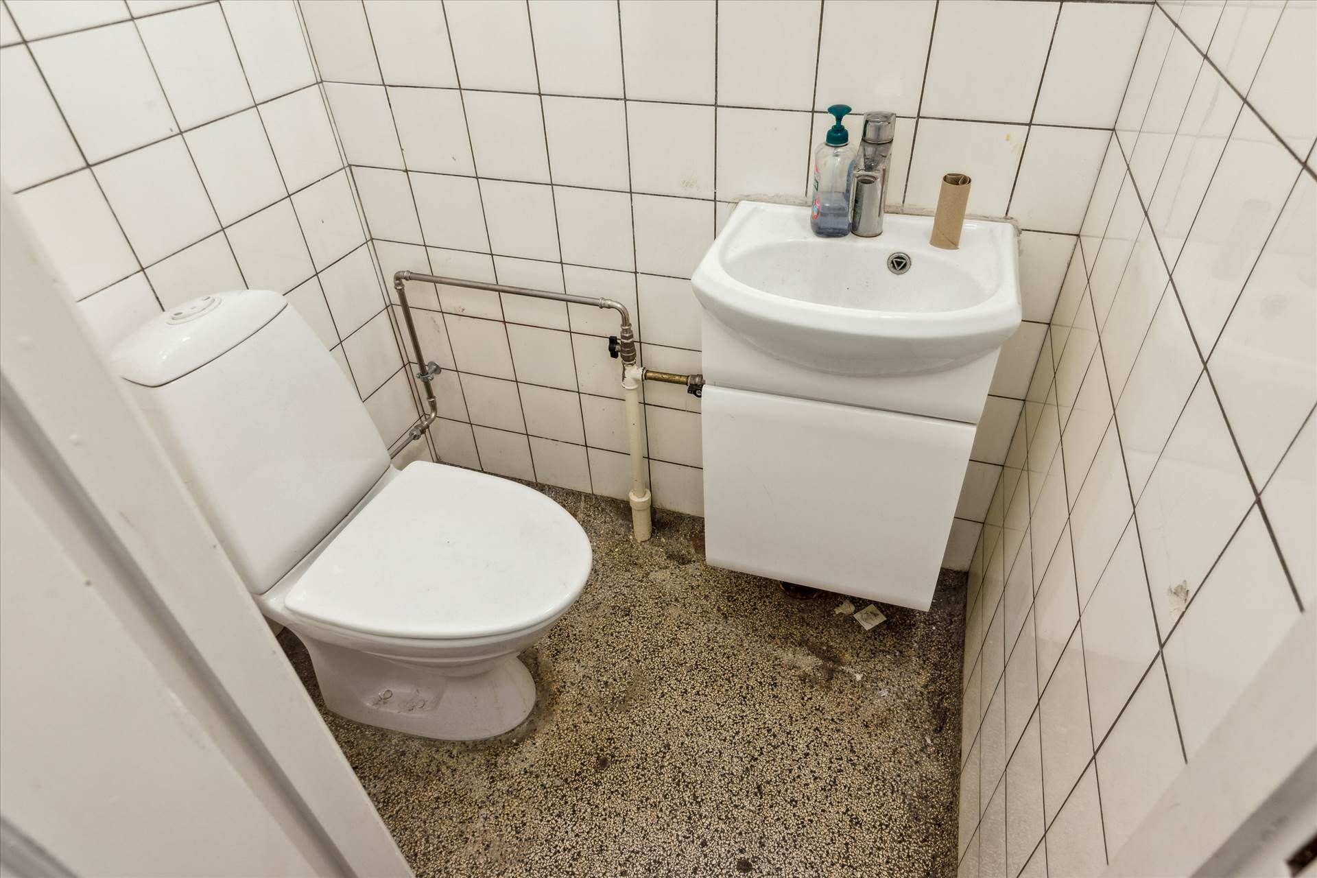Et lille, hvidt badeværelse med toilet, hvidt badekar og hvidt vask. Under vasken er der en hvid skab og en håndsæbe