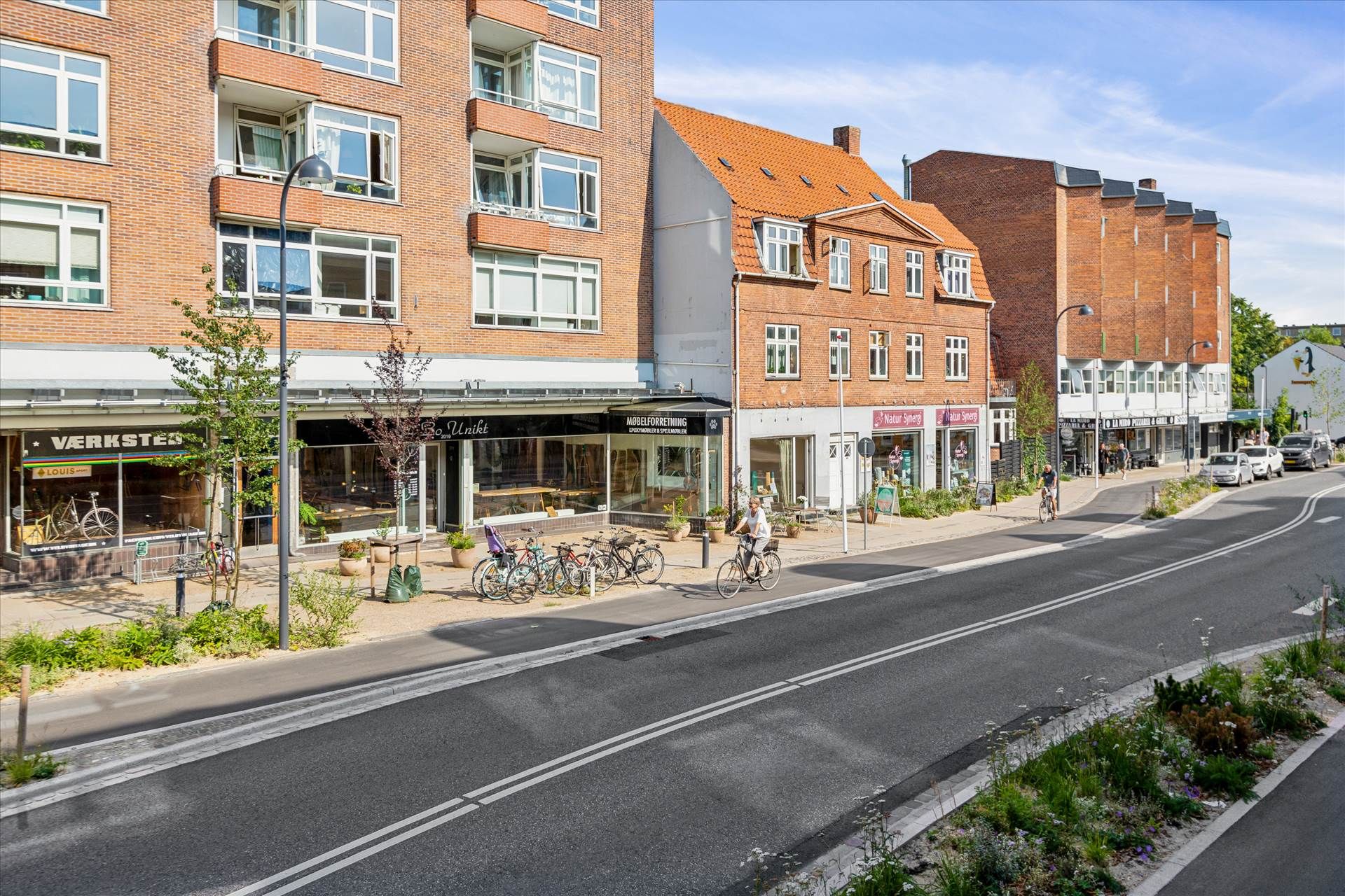 En tosporet asfaltvej med grønne planter og blomster langs siderne, hvor der er to cyklister, der kører langs med biler på