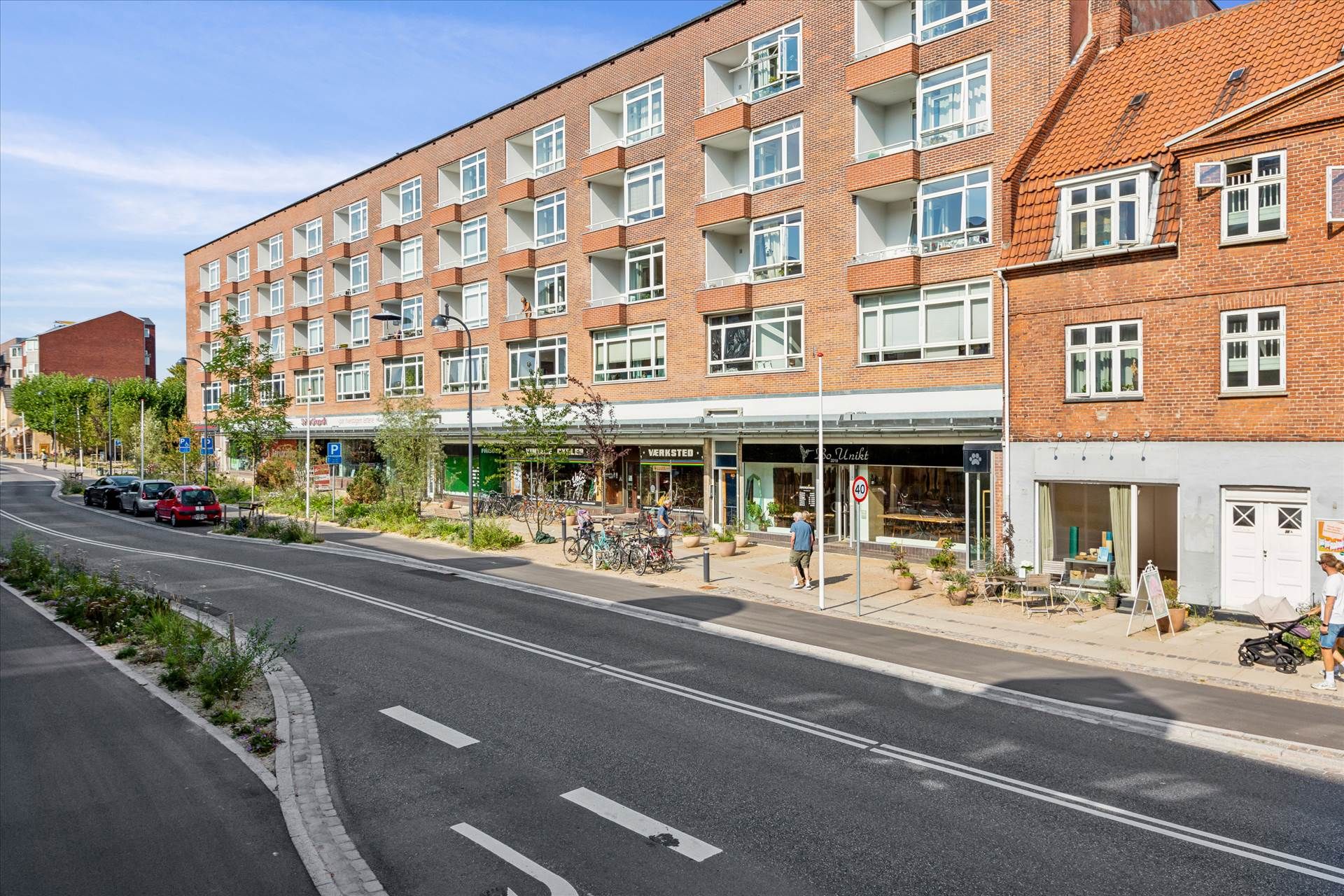 Butikslokaler i Søborg med parkeringspladser og grønne planter langs vejen og to fodgængere med en barnevogn