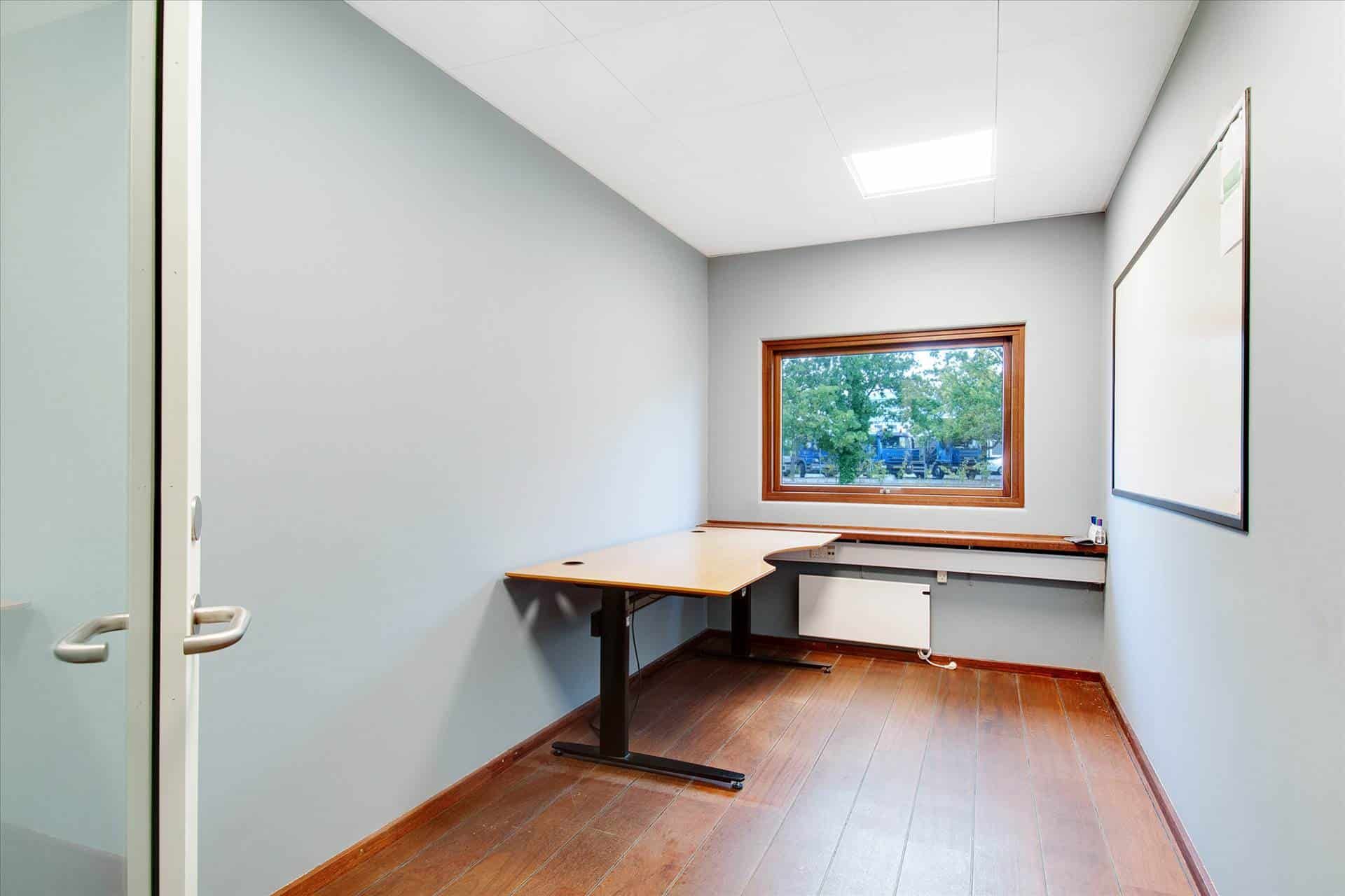 Kontorrum med højt til loftet, lysevinduer og trægulve. Der er et bord og en whiteboard på væggen.