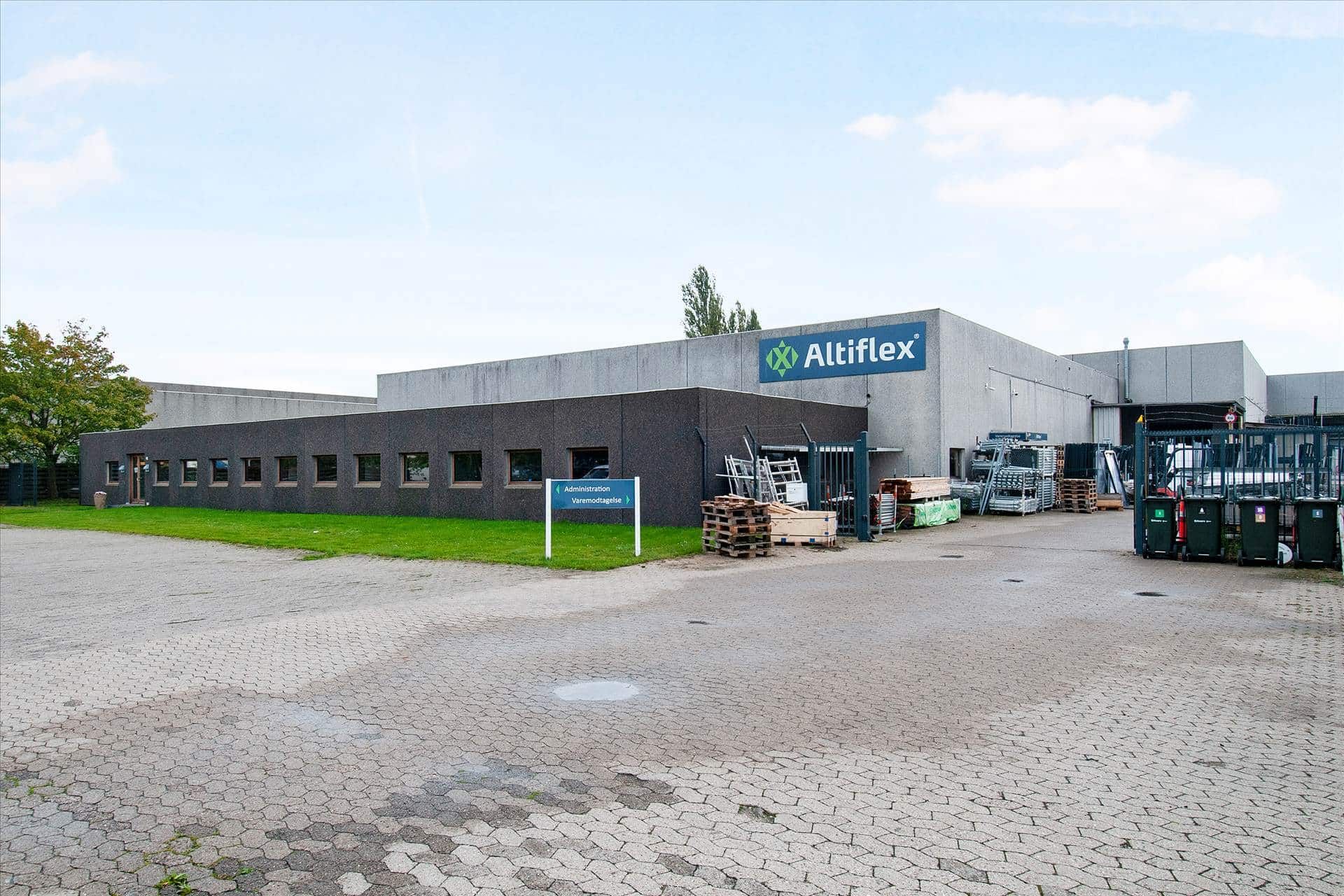 Kontorbygning med logoet Altiflex. En parkeringsplads med grønne søjler og to papirkurve. Administrationsskilt udenfor