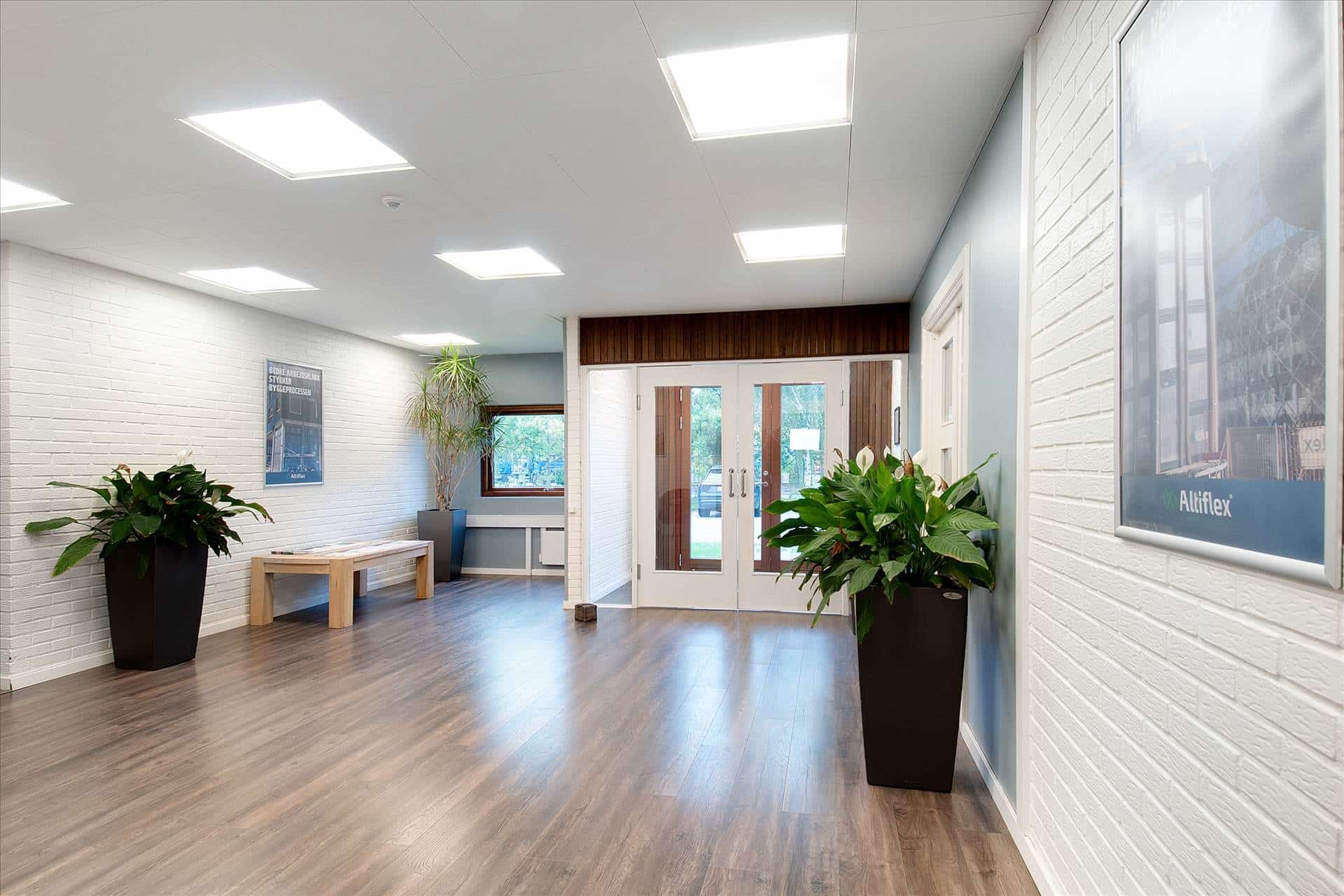 En stor lobby med hvide vægge og lyse loftslamper samt større planter i sort. Der er også et bord og et vinduesparti med