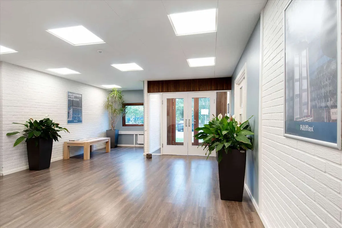 En stor lobby med hvide vægge og lyse loftslamper samt større planter i sort. Der er også et bord og et vinduesparti med