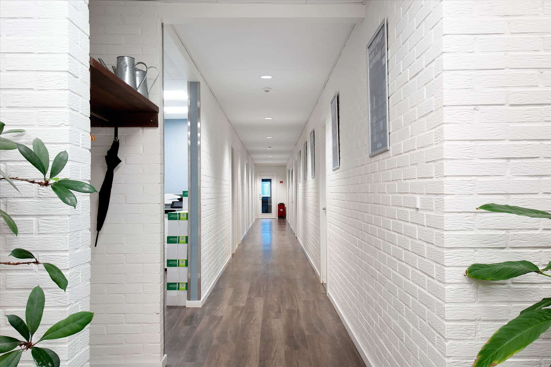 En lang hal med hvide vægge og lysegrå gulv. Der er et sæt plantager og et stel med tøj ved siden af en dør. Der er også et