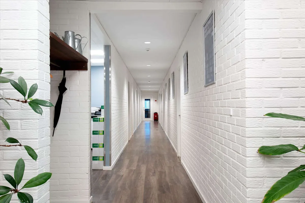 En lang hal med hvide vægge og lysegrå gulv. Der er et sæt plantager og et stel med tøj ved siden af en dør. Der er også et