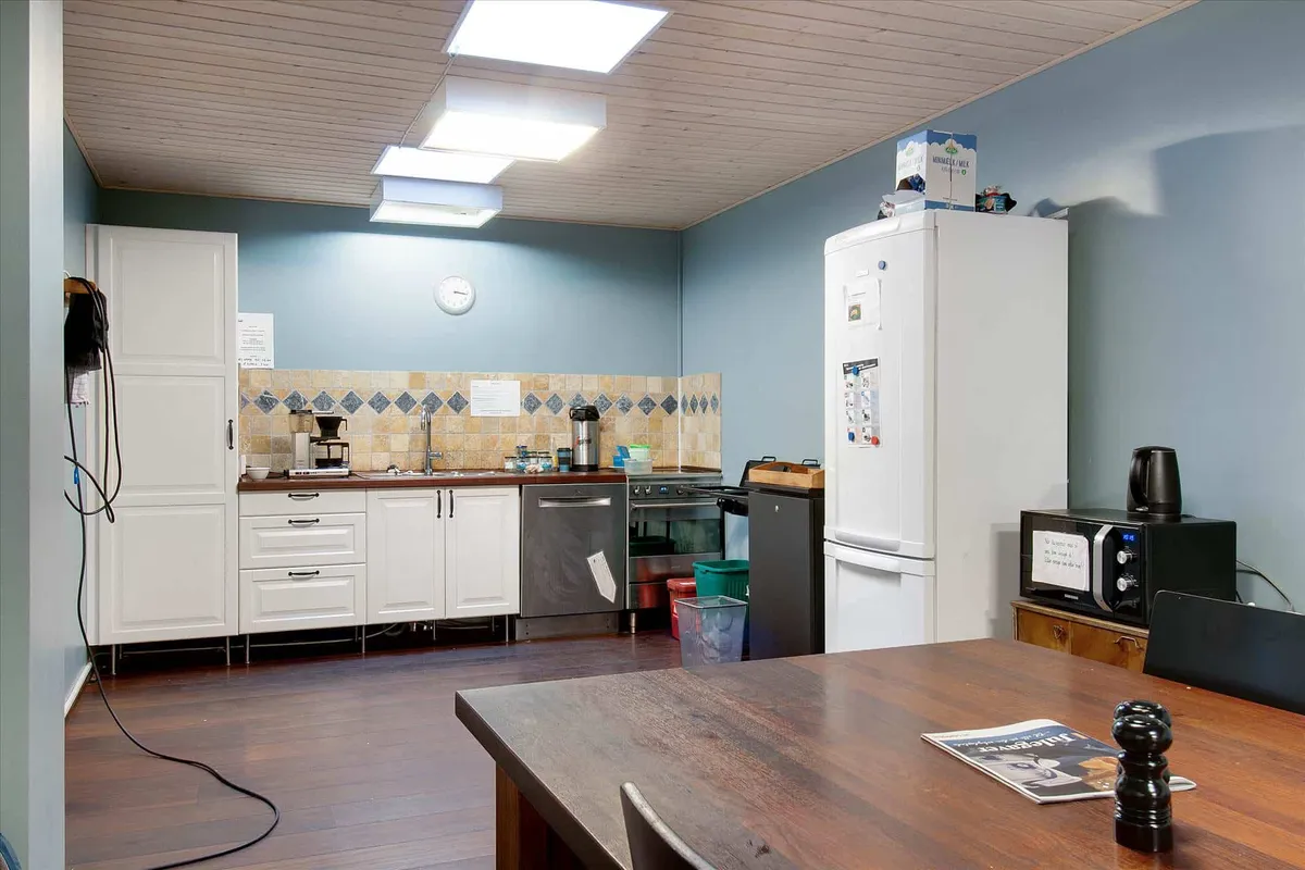 Inde i et moderne køkken er der en stor hvid køleskab, en mikrobølgeovn, og en træbord med en avis og en salt- og peberkværn