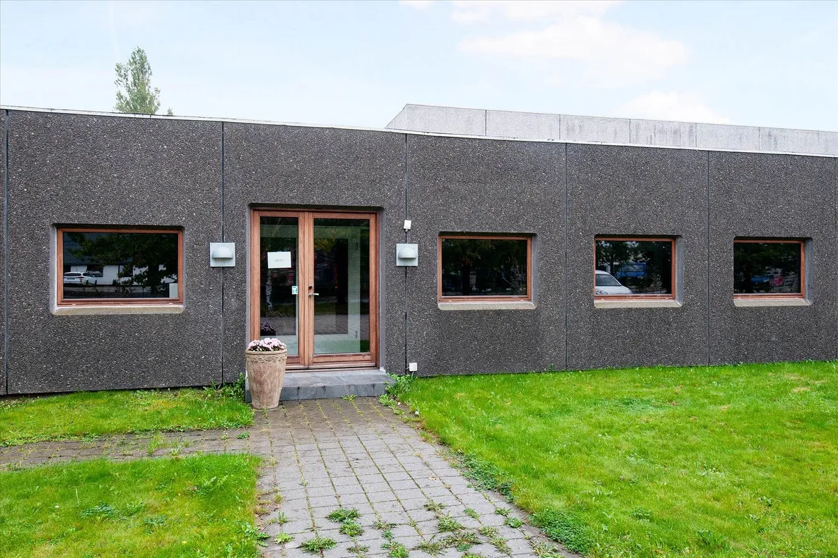 Et moderne kontorbyggeri med en stor glasdør og fire vinduer med brunt indramning og en stor potte med blomster foran