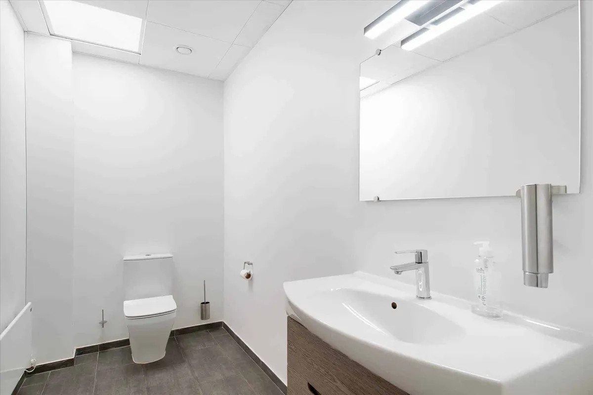 En hvidt malet badeværelse med lysekroner, en stor spejl, et toilet, en håndvask og en håndsæbe dispenser