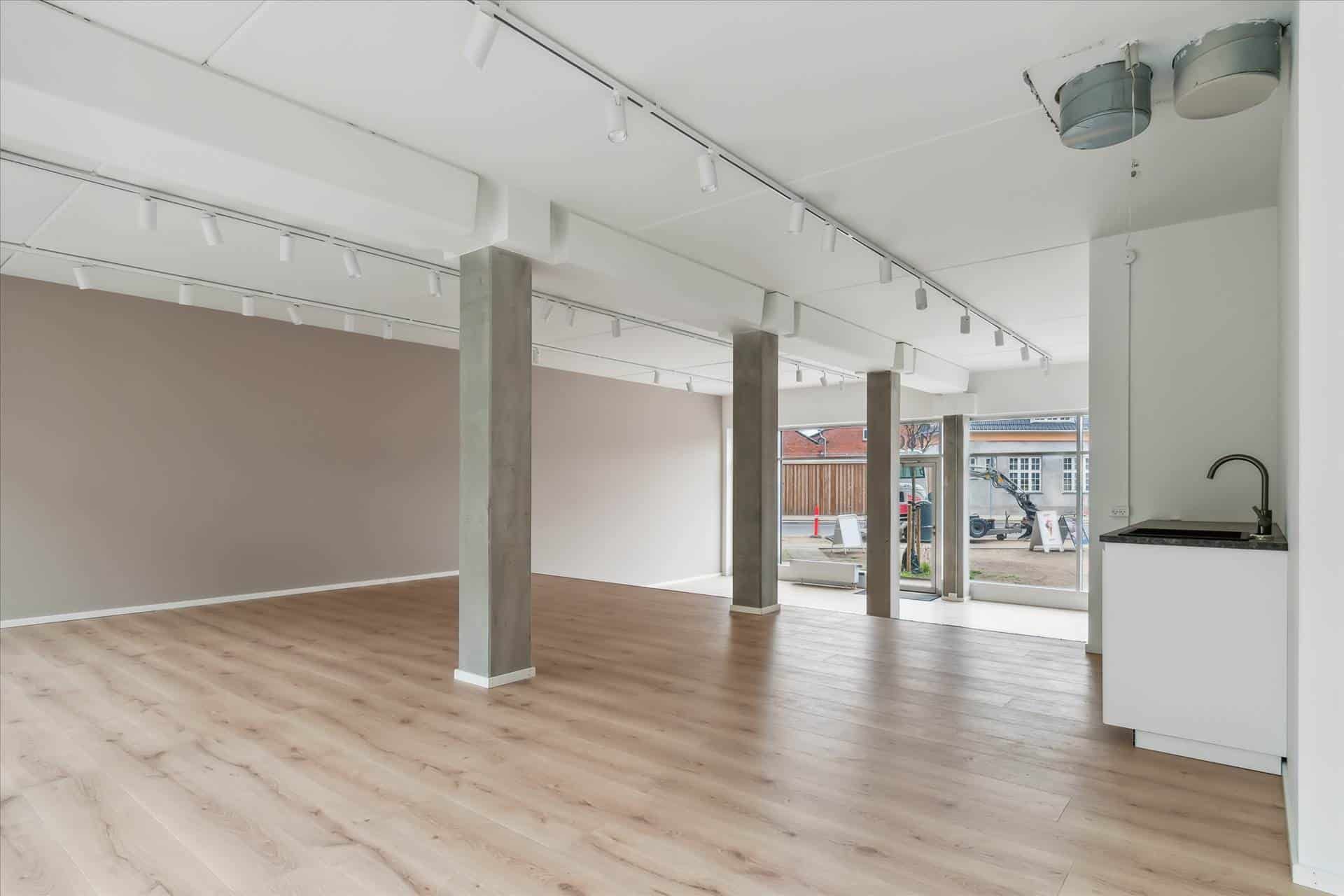 Et stort, tomt rum med lyse trægulve, hvide vægge, højloft og flere støtter. Et glasdør fører ud til byggeområdet.