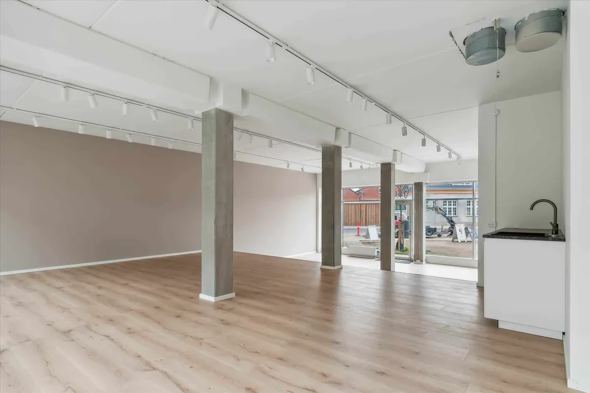Et stort, tomt rum med lyse trægulve, hvide vægge, højloft og flere støtter. Et glasdør fører ud til byggeområdet.