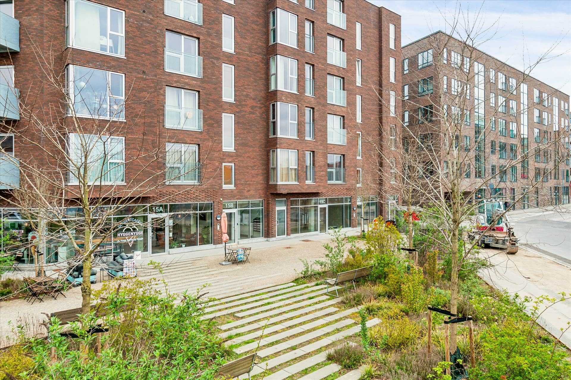 To etager højt bygning med facader af rød mursten og glasvinduer. Et nyt showroom er under opførelse med buske og træer. En