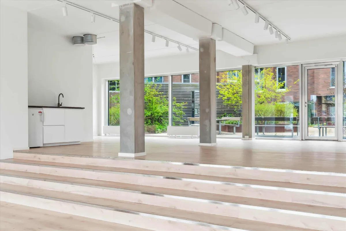 Et rum med høje lofter, hvide vægge, tre kolonner og lyse trægulve, hvor der er åbne ruder til have med grønne planter og et