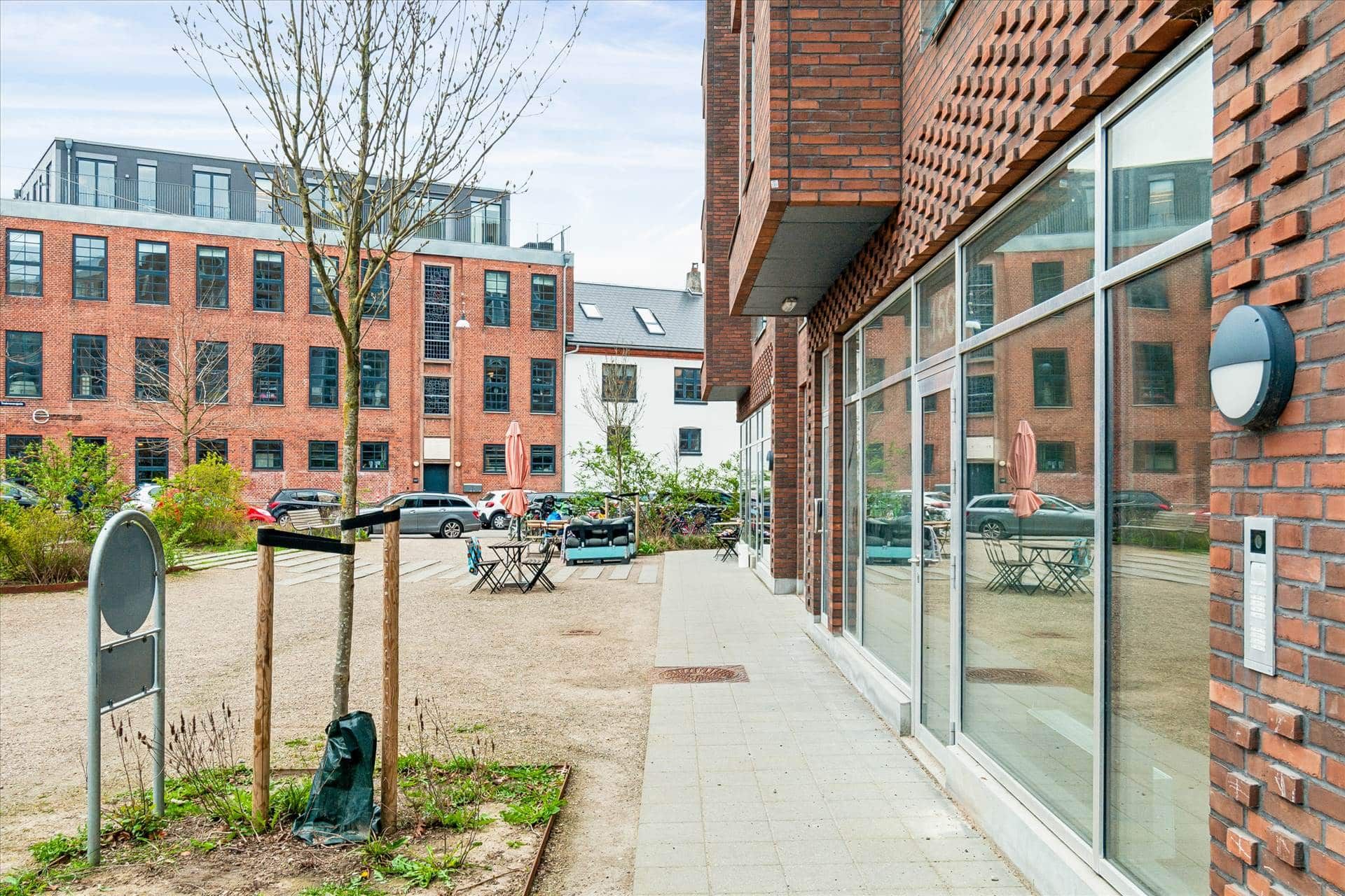Moderne bygning med glas og murstensfacade, udendørs cafeområde, grønne planter og bord med paraplyer, lyseblå himmel og