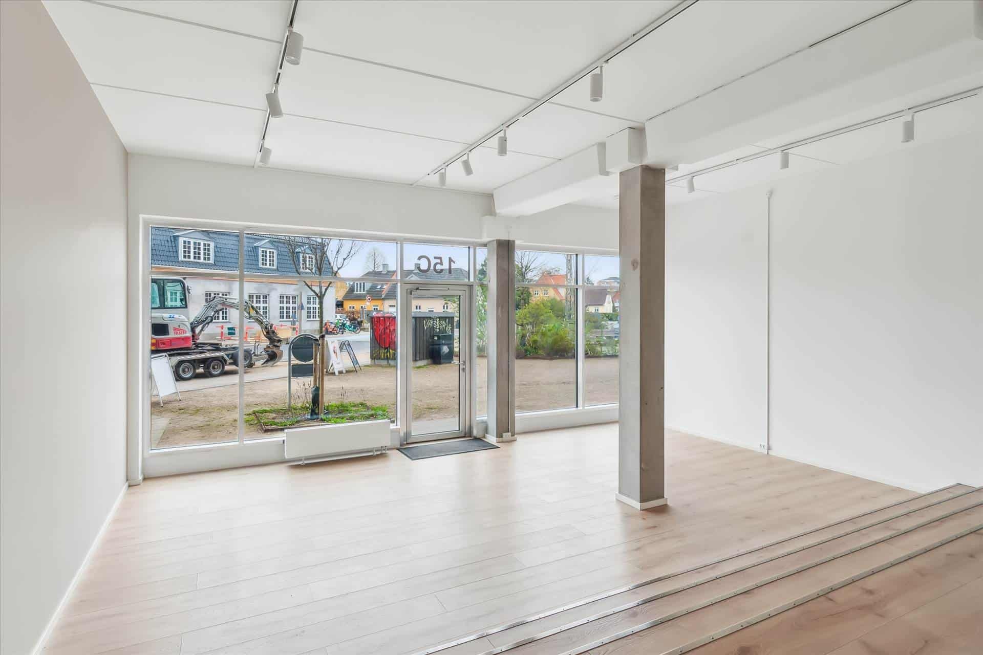 Showroom med glasvinduer og lyse loftslamper, hvor man ser en ekskavaator og en bygning med blå tag på udenfor