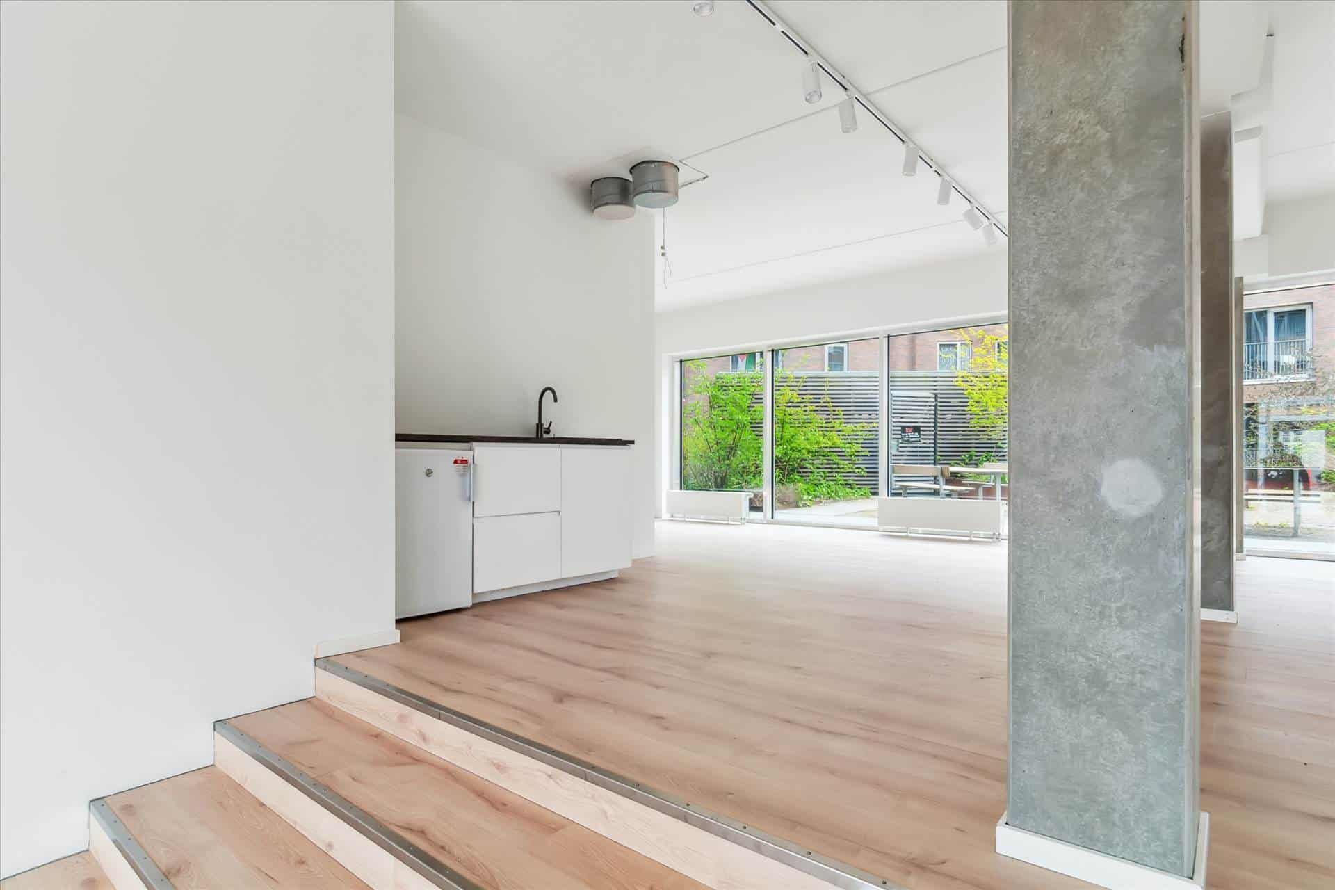 Moderne kontorlokaler med højloft og lyse vindueskarme, hvide vægge, trægulv og en minimalistisk køkkenenhed med håndvask og