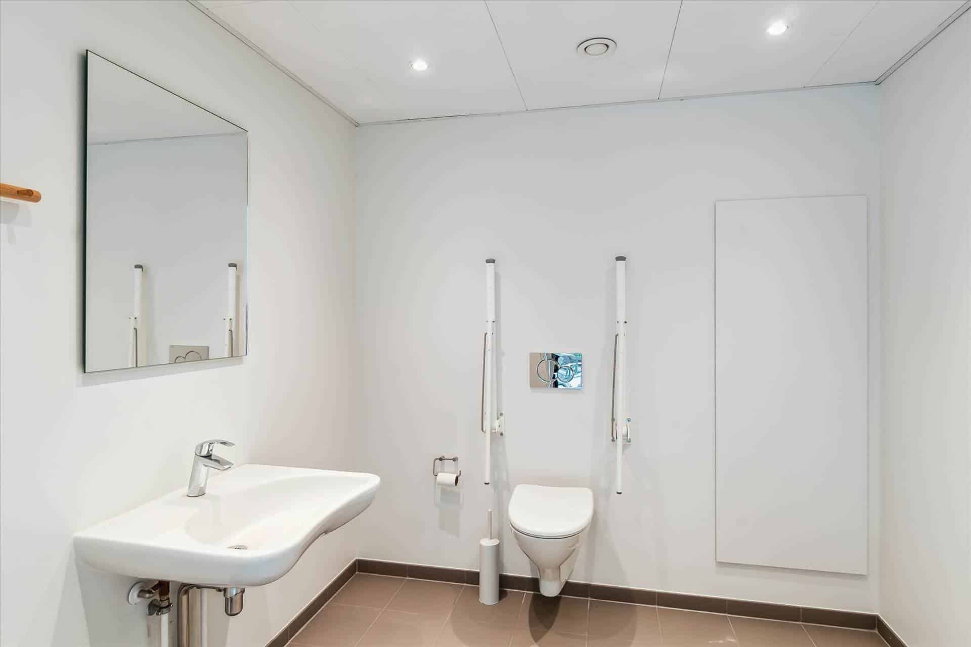 Alt tekst: En minimalistisk badeværelse med hvid keramik, moderne spejl og hvid toilet med støtter og gul gulv.