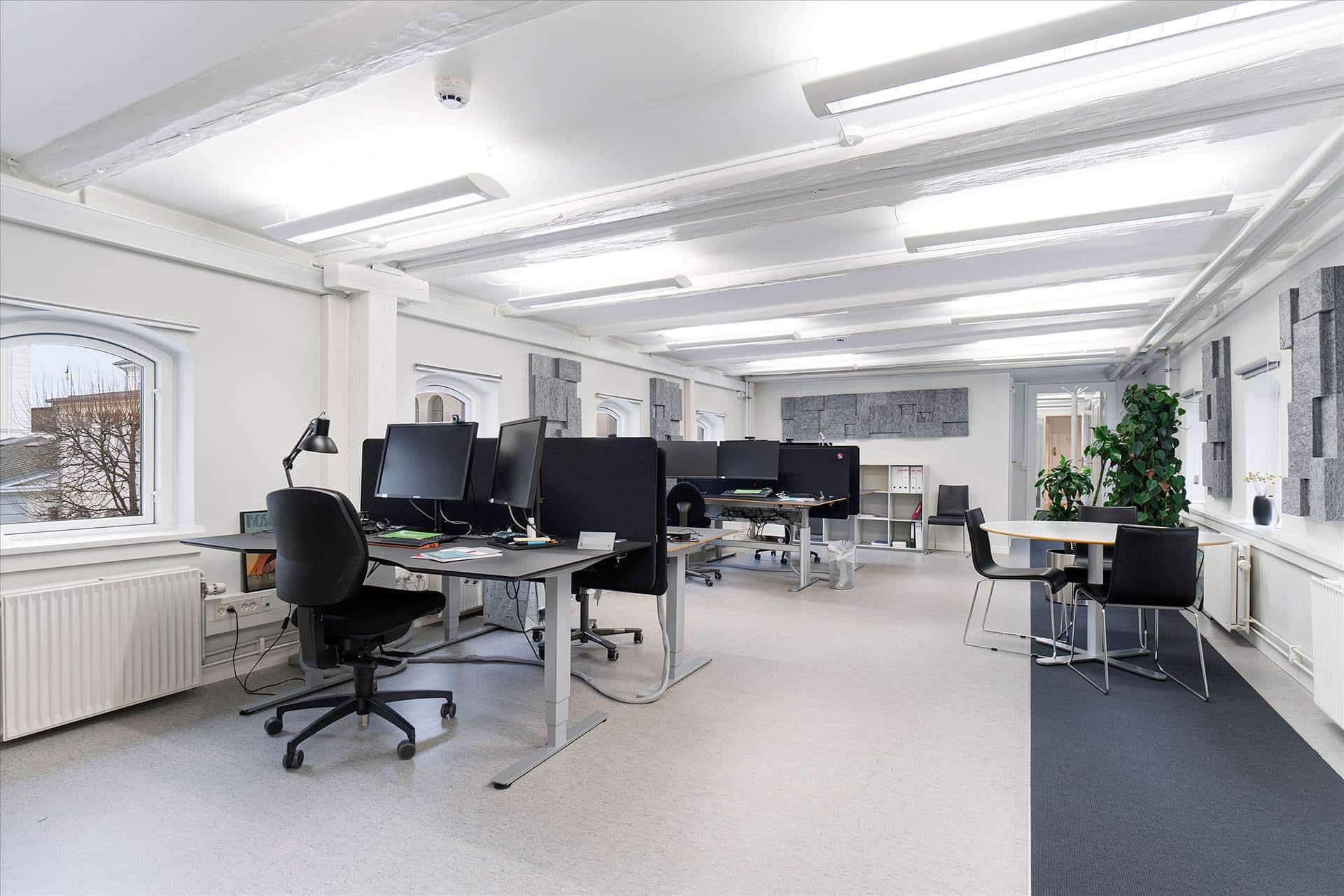 Kontor med skriveborde, stole, skrivebordslamper, computere og et rundt bord med stole. Et større sted med højt lys og højt