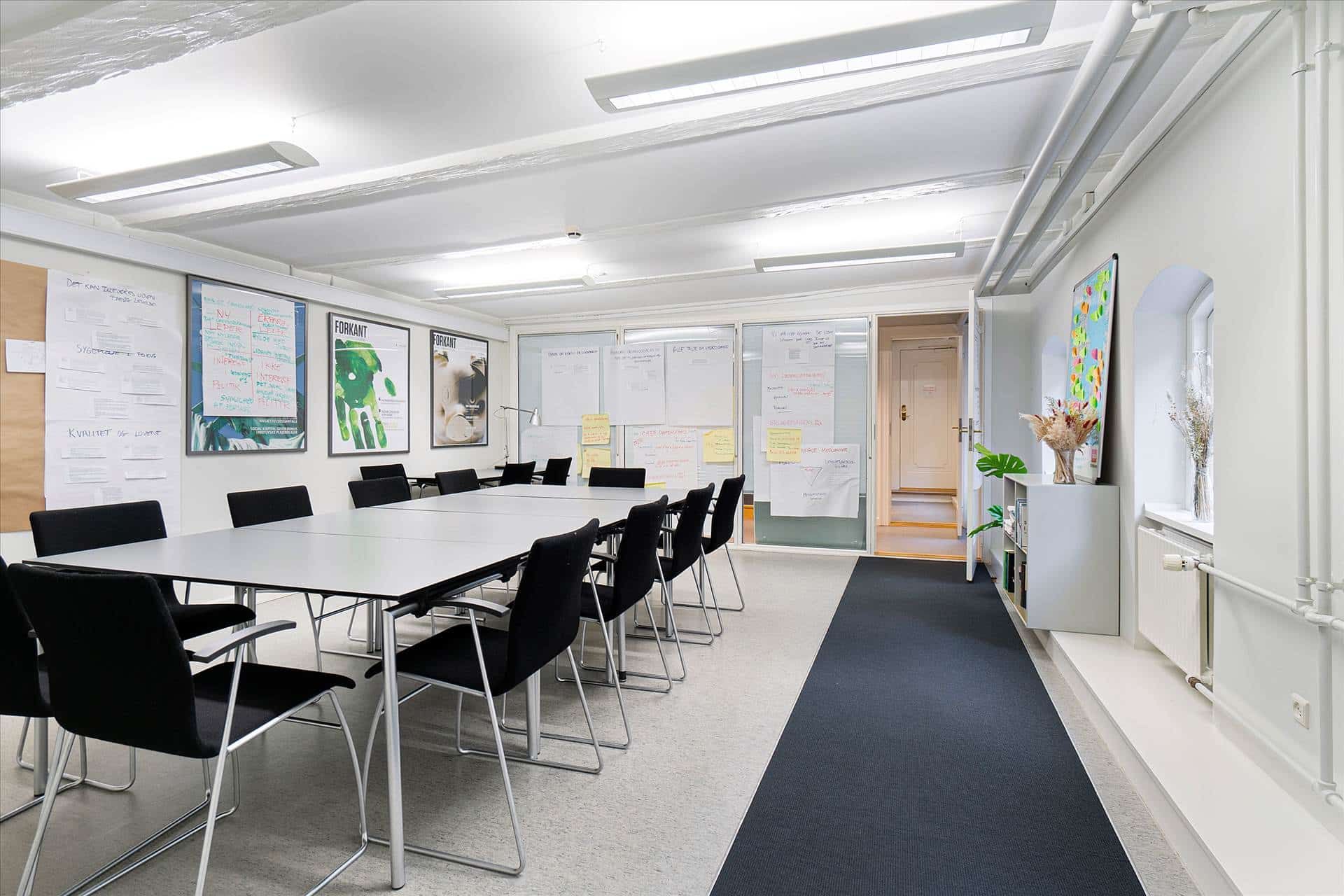 Mødelokale med hvide vægge, en lang bord og stole, og lyse skrifttavler og plakater med forskellige informationer og
