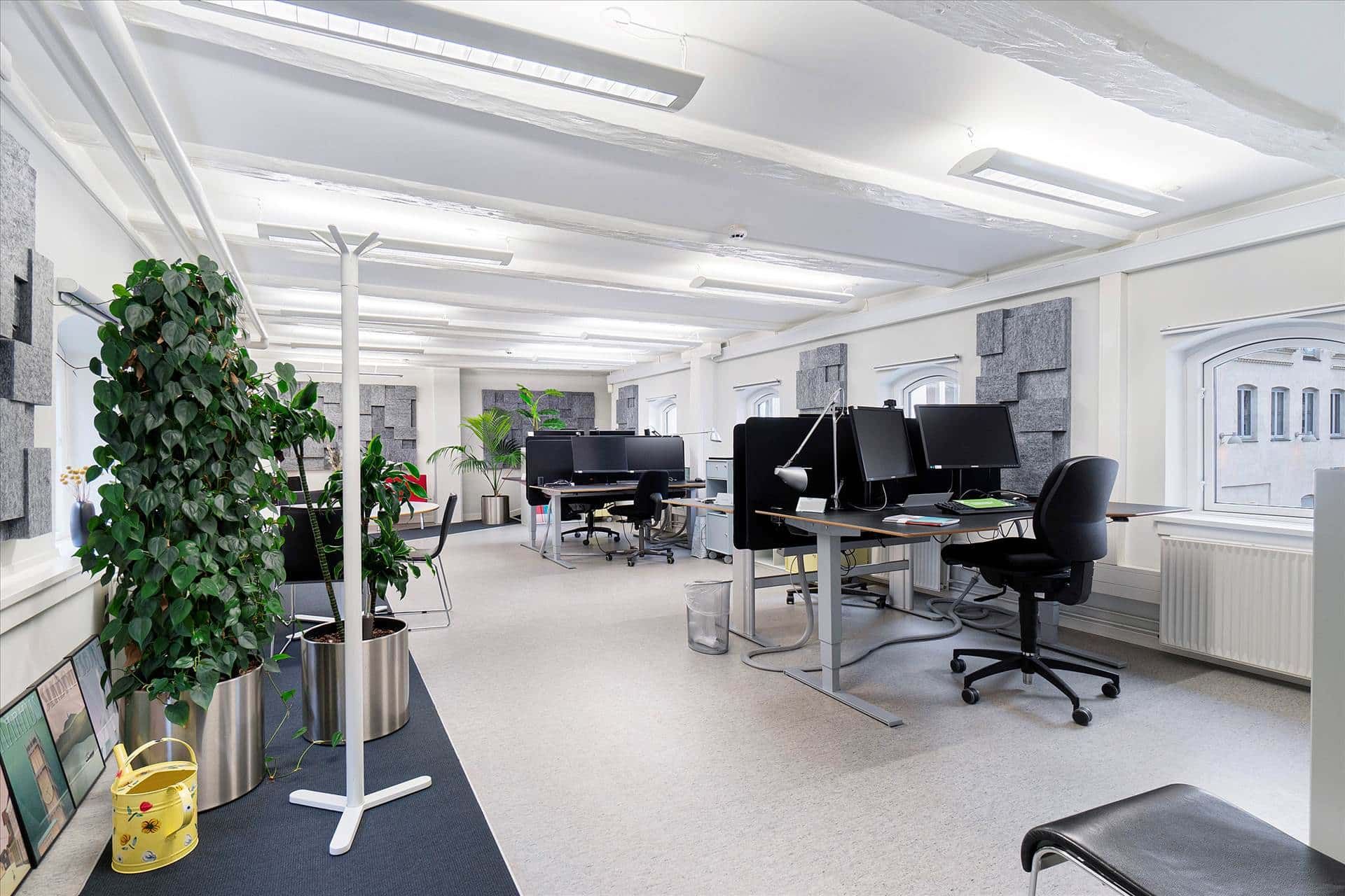 Moderne kontor med skriveborde, stole, plante og højt gulv. Loft med lysekroner og vindue med udsigt til bygning.