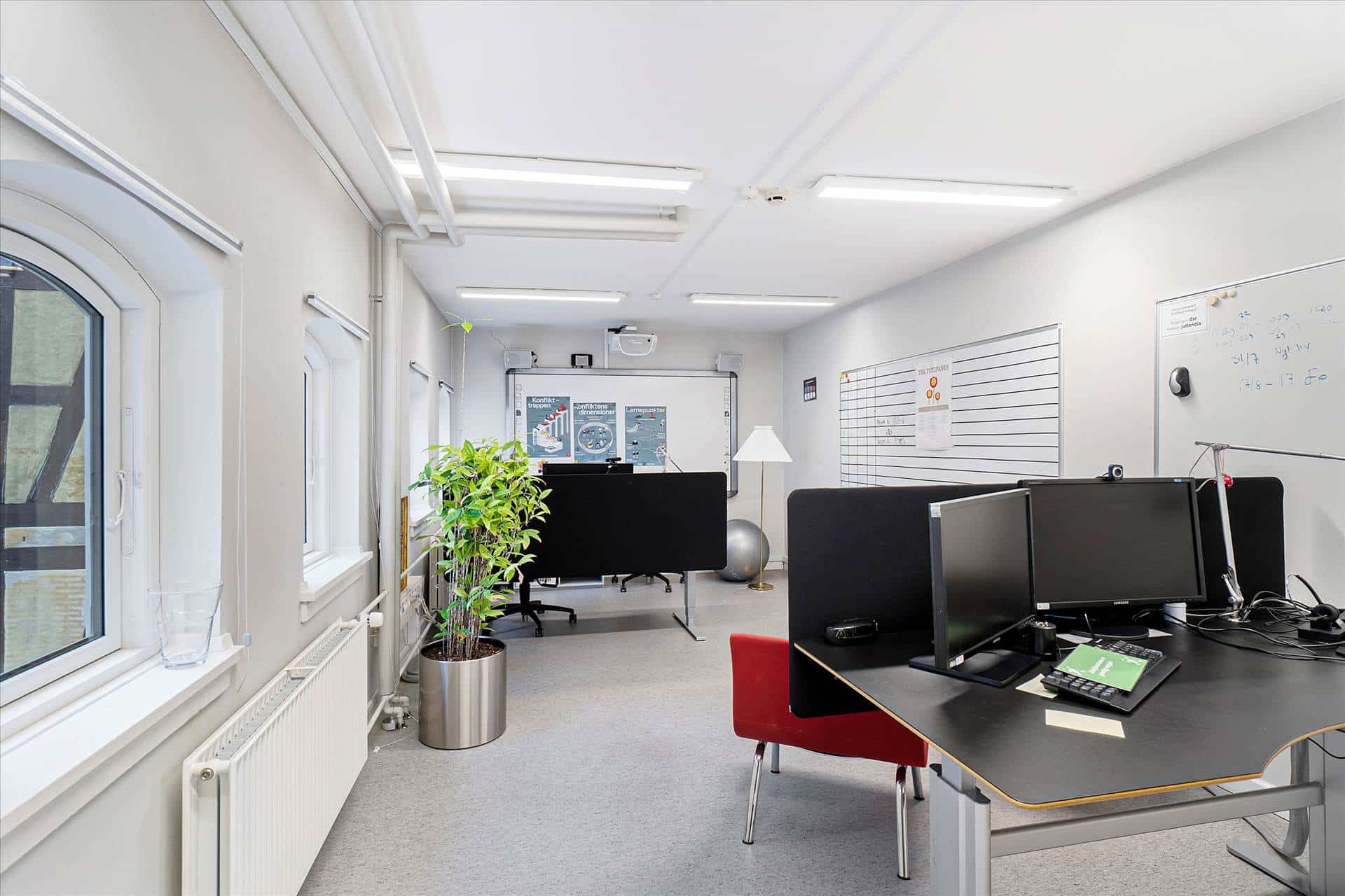 Ledig kontor med skrivebord, computer, to skærme, rødt stol og lampe samt plante og kalender på væggen